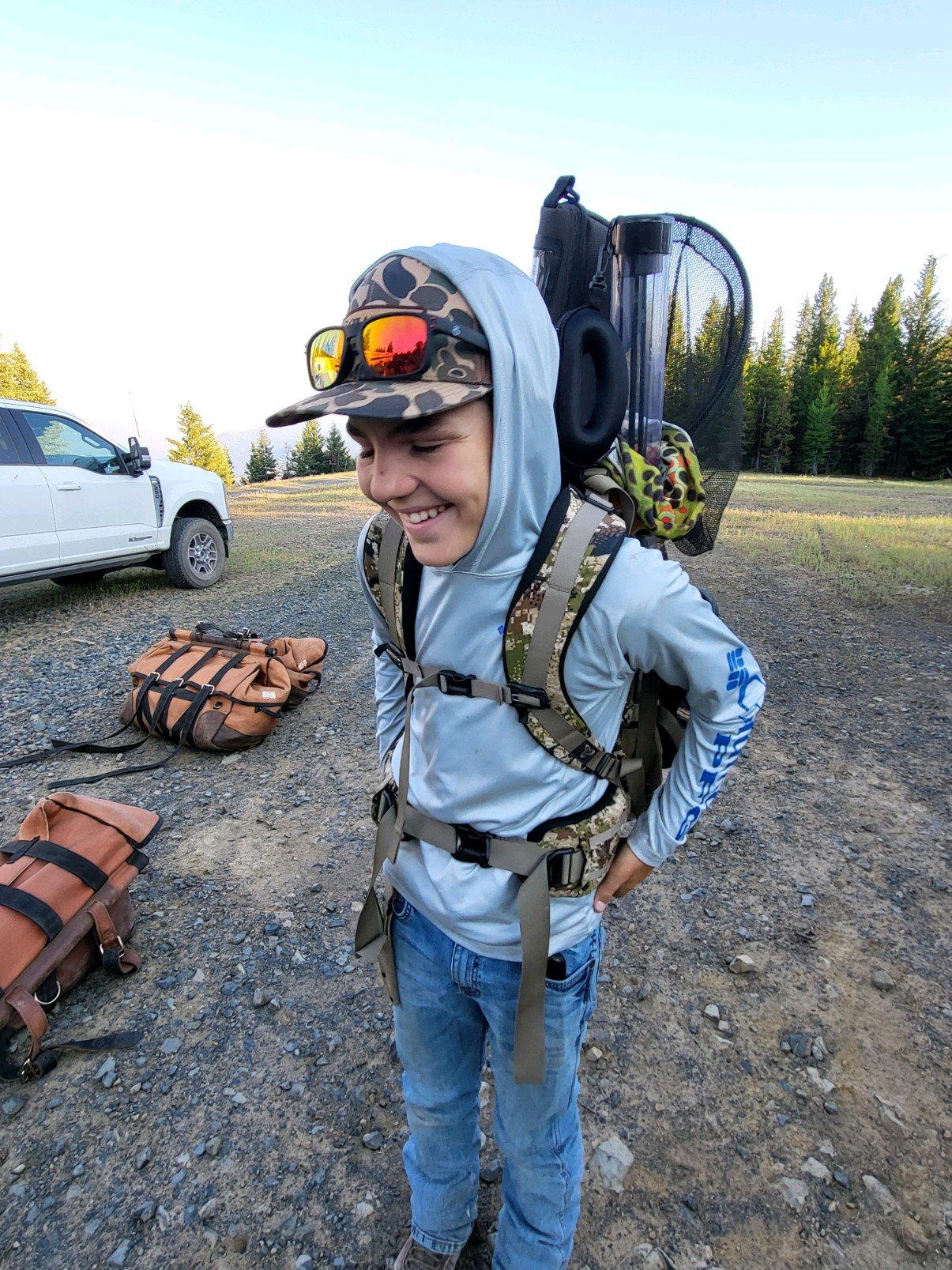 Young man with a backpack, sunglasses, and a camouflage cap, standing outdoors on a gravel field with trees and a white vehicle in the background, smiling.