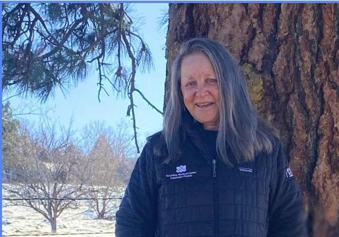 An older woman outdoors standing next to a large tree with a blue sky in the background, wearing a black Patagonia jacket and smiling.