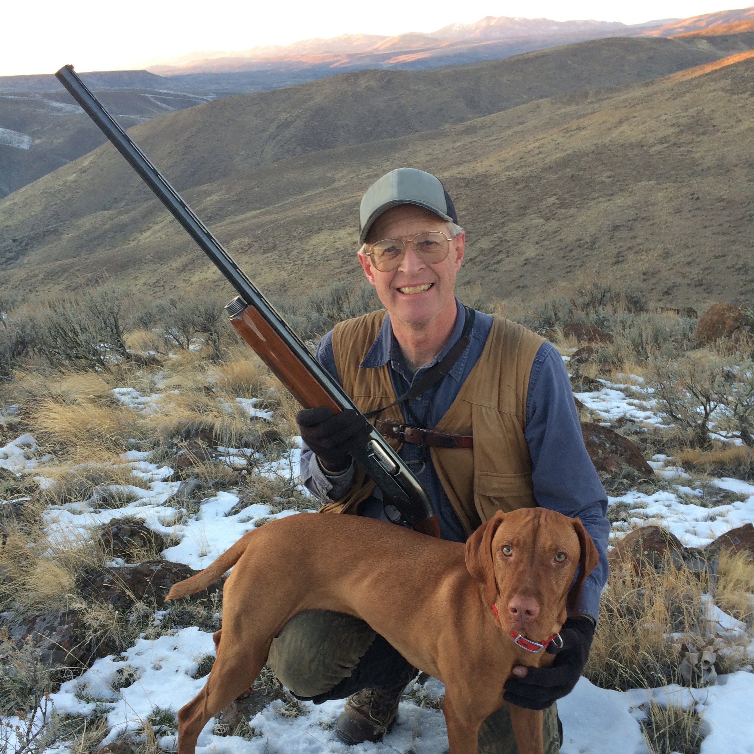 A man in outdoor clothing holding a shotgun and smiling, kneeling with a brown Labrador Retriever in a snowy, mountainous landscape.