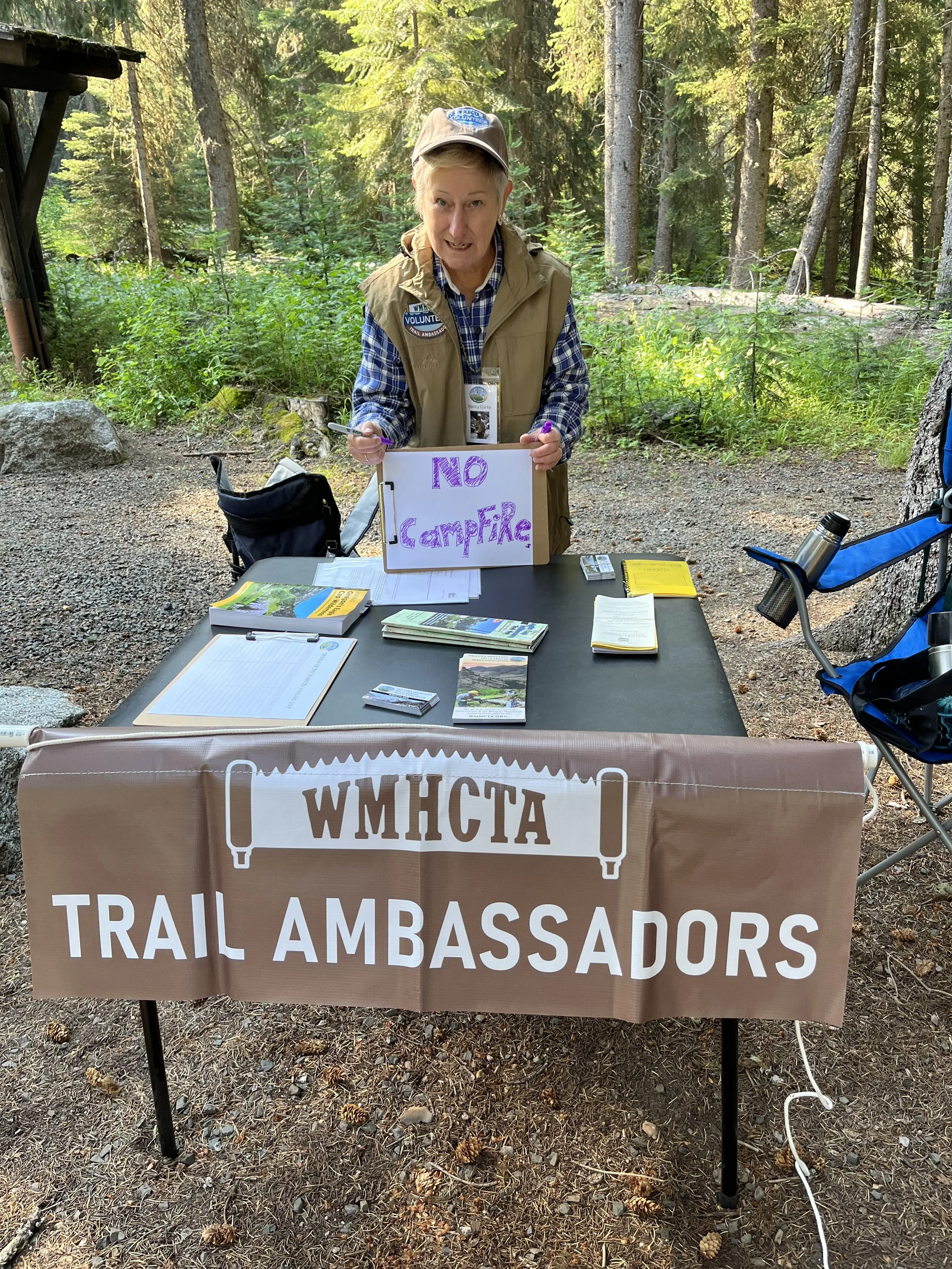 A woman standing at a trailhead table outdoors in a forest, holding a sign that reads 'No Campfire'. The table has brochures, papers, and a yellow book, with a large brown sign reading 'WMHCTA Trail Ambassadors' hanging from the front.
