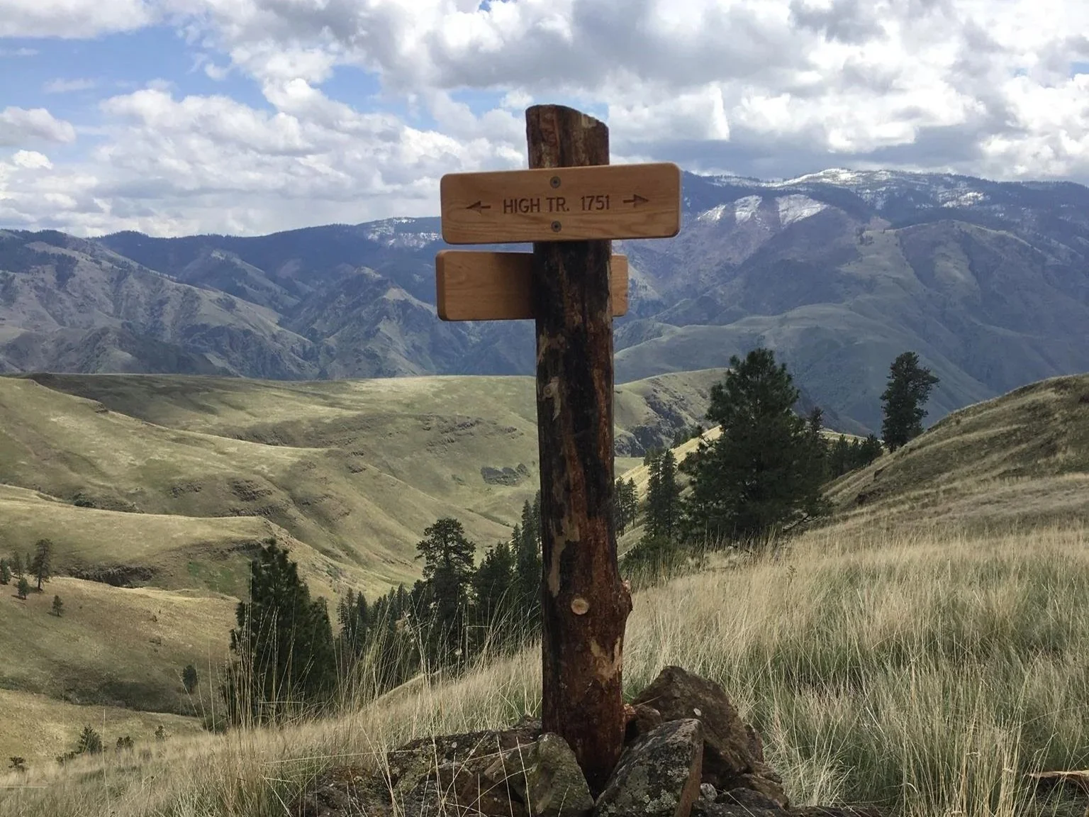A wooden trail sign in a mountainous landscape, indicating 'High Tr 1751' with arrows pointing in both directions, surrounded by grassy hills and trees under a cloudy sky.