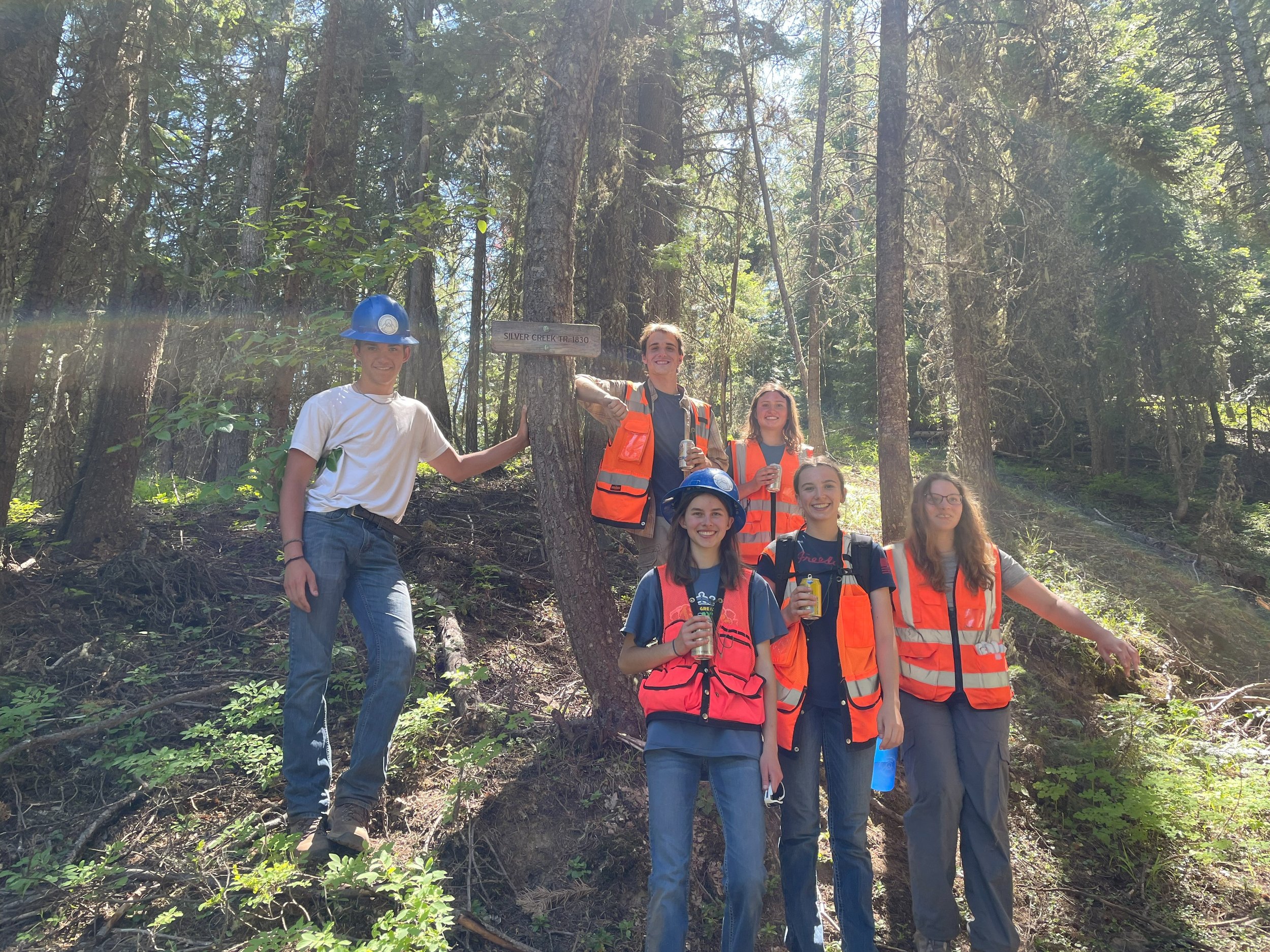Group of six young adults outdoors in a forest, wearing safety vests and helmets, some holding drinks, smiling for a photo near a sign on a tree that reads 'Silver Creek Trail 1830'.