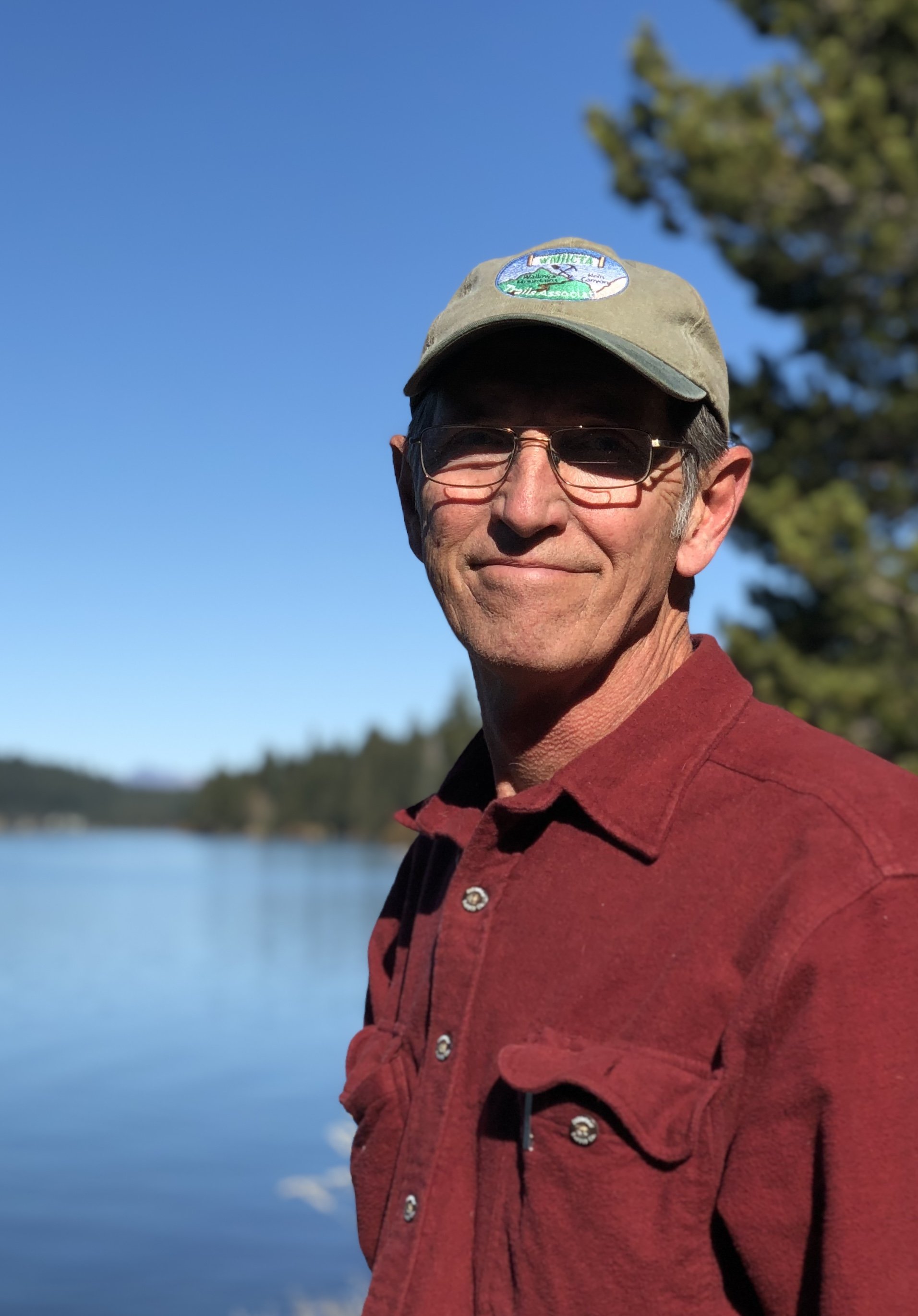 A middle-aged man wearing glasses, a beige cap, and a red shirt standing outdoors near a body of water, with a blue sky and trees in the background.