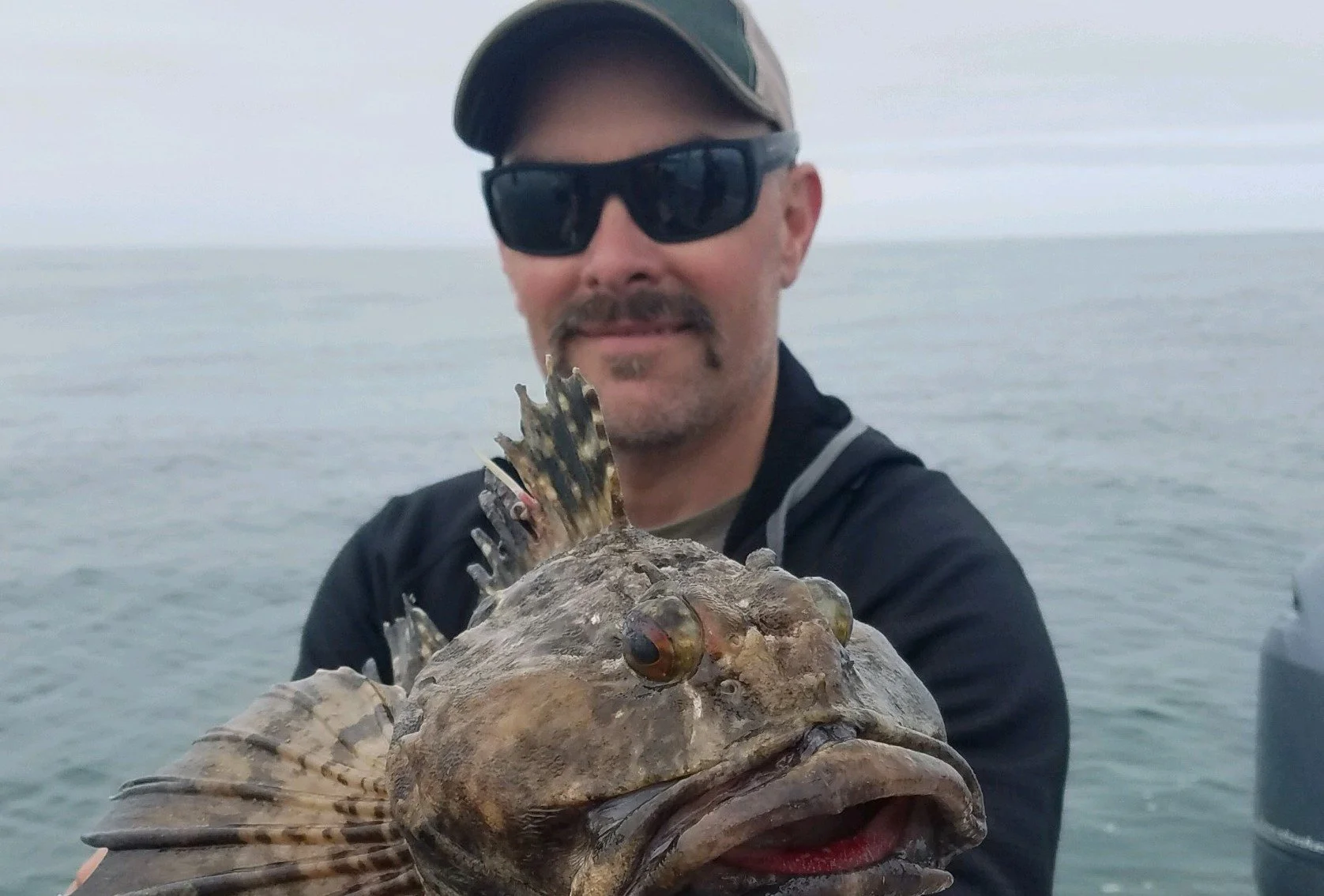 Man wearing sunglasses and a baseball cap holding a large fish with spiky fins on a boat in the ocean.