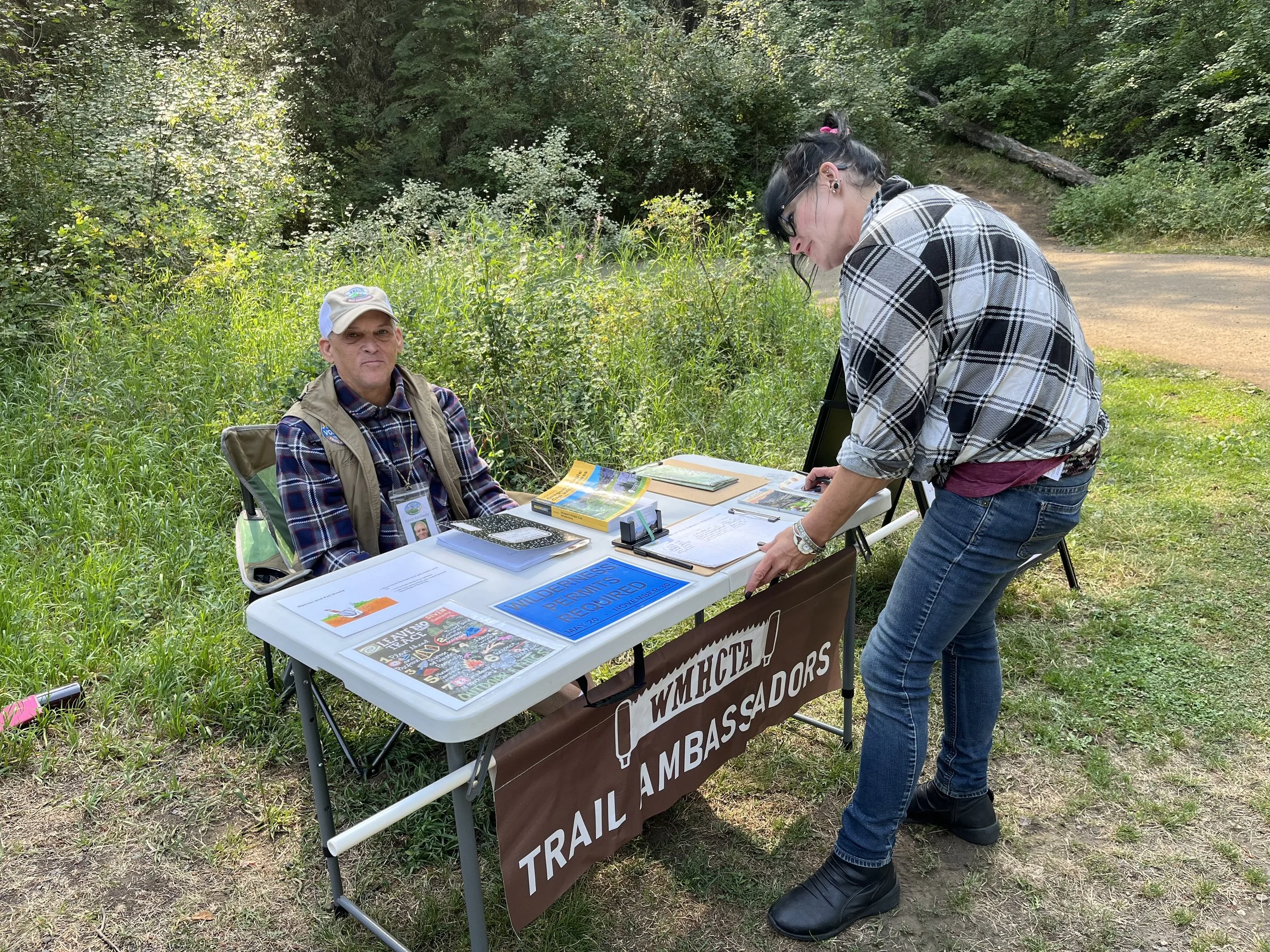 A woman standing at a table with trail and outreach signs, speaking with an older man seated behind the table in outdoor greenery.
