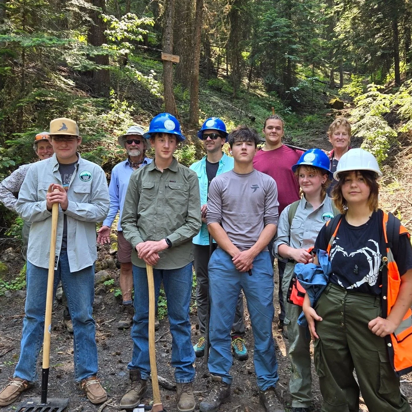 Group of people in a forest, some wearing hard hats, holding tools, and dressed in outdoor gear, suggesting they are on a hiking or outdoor work trip.