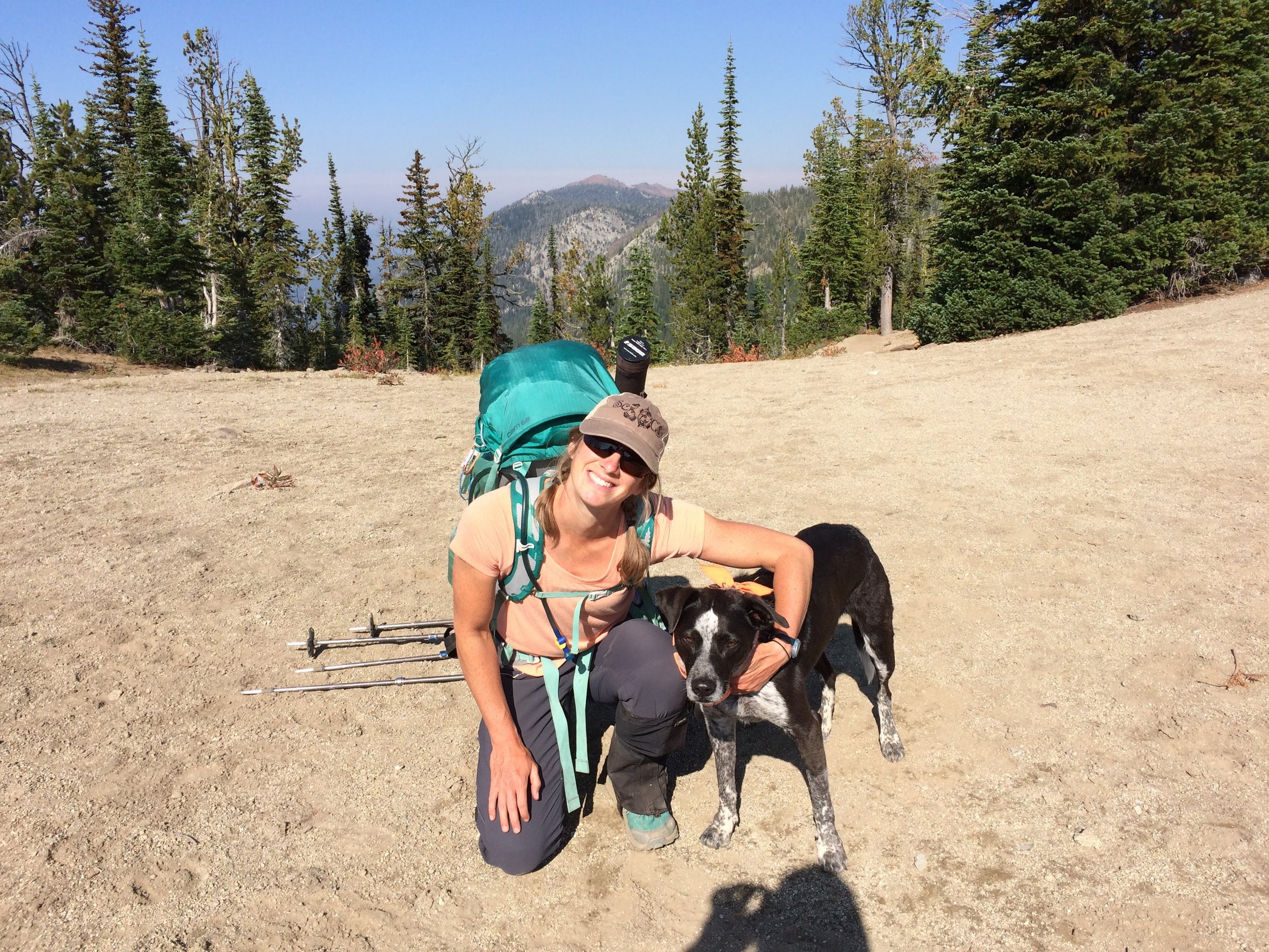 A woman with a backpack and hiking gear kneeling next to a black and white dog on a mountain trail with trees and mountains in the background.