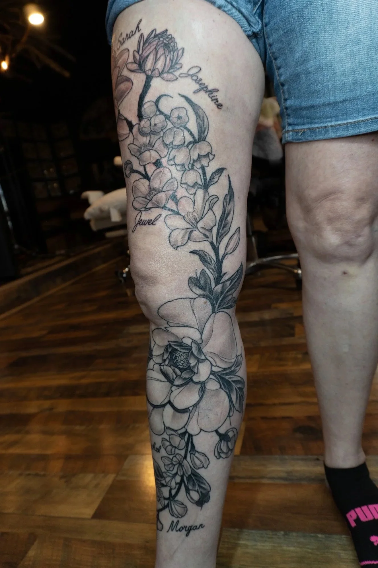 A person's leg with a detailed black and gray floral tattoo, including roses and other flowers, with the names Sarah, Josephine, Jewel, and Morgan written near the flowers.
