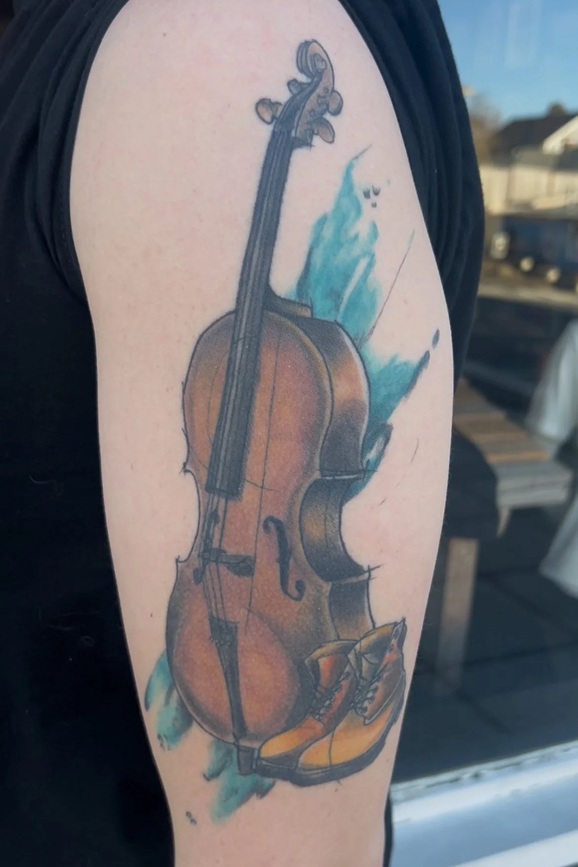 Tattoo of a violin, bow, and a pair of yellow shoes on a person's upper arm.