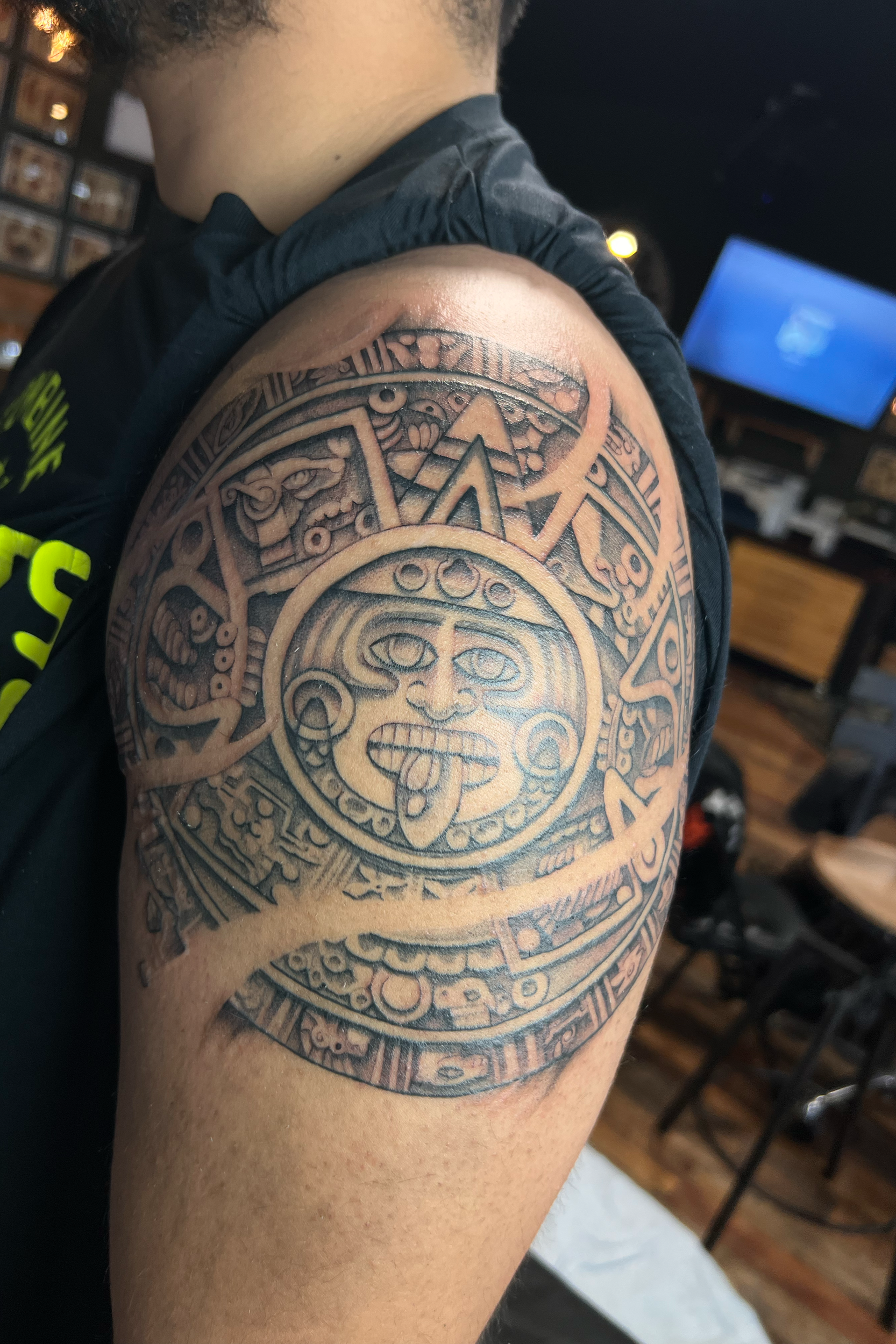 A person with a large Aztec-style tattoo on their upper arm and shoulder. The tattoo includes traditional Aztec symbols and a central face with a tongue sticking out.