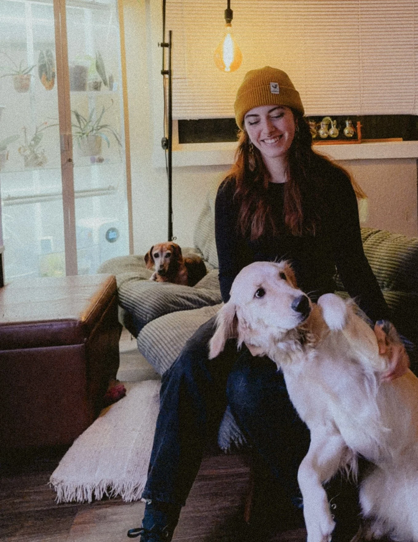A woman wearing a yellow beanie sitting on a couch with two dogs, one golden retriever and a smaller dog in the background, in a cozy living room.