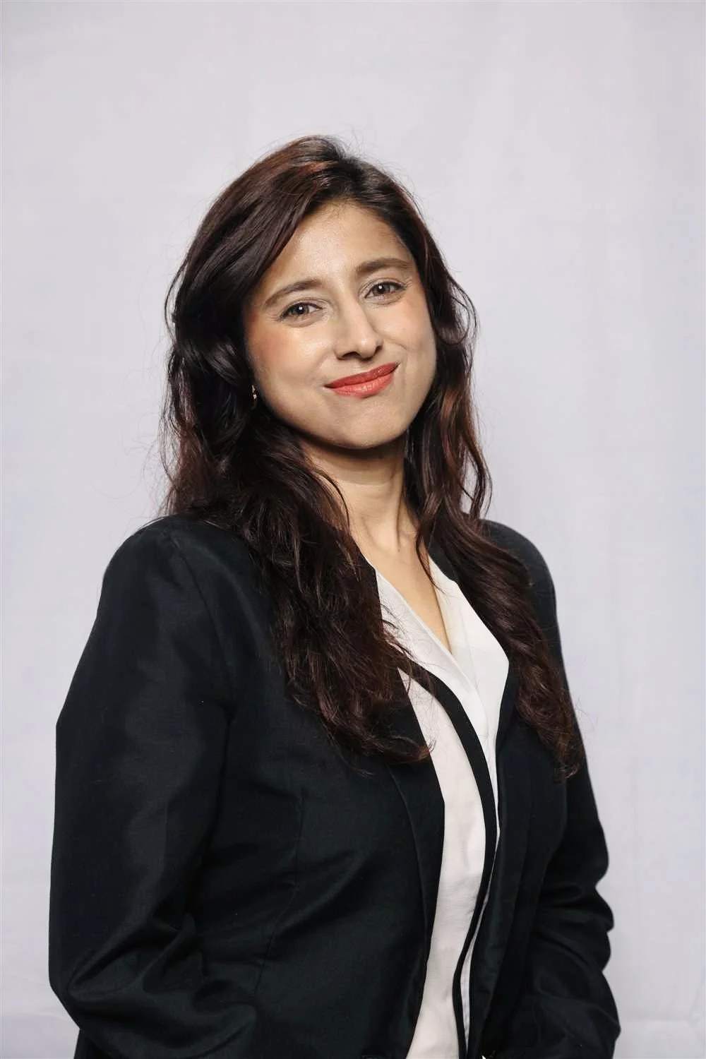 A woman with brown, wavy hair and light skin, wearing a black blazer over a white blouse, smiling against a plain white background.