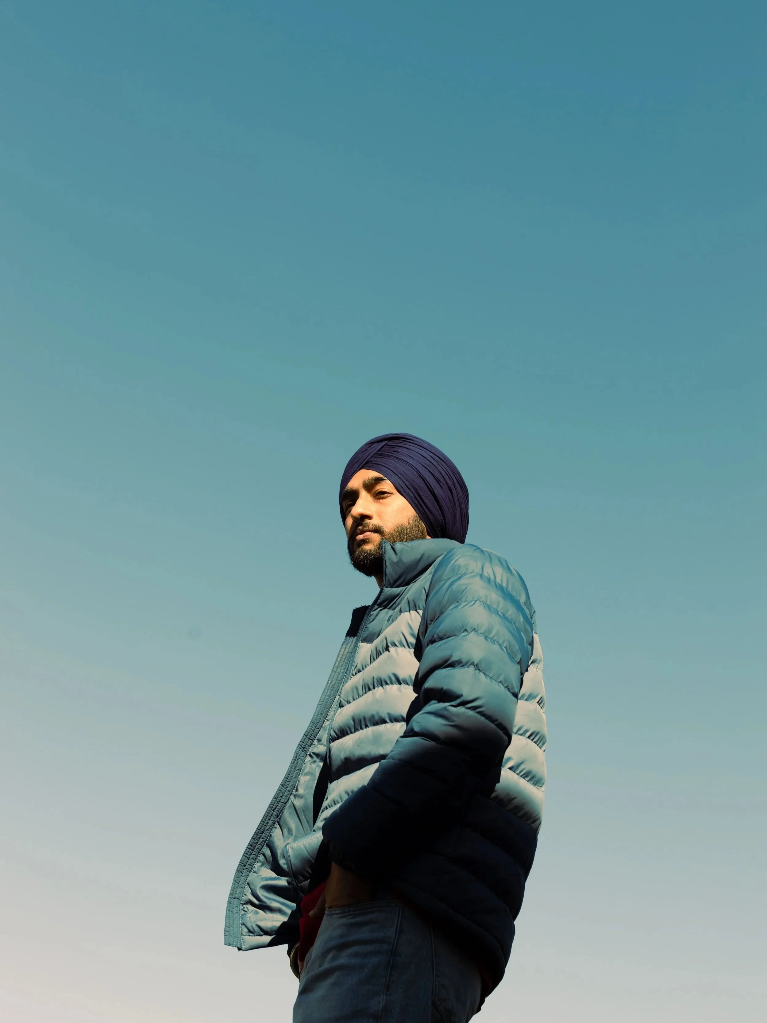 A man wearing a dark blue turban and a light blue and black puffer jacket stands outdoors against a clear sky, looking into the distance.