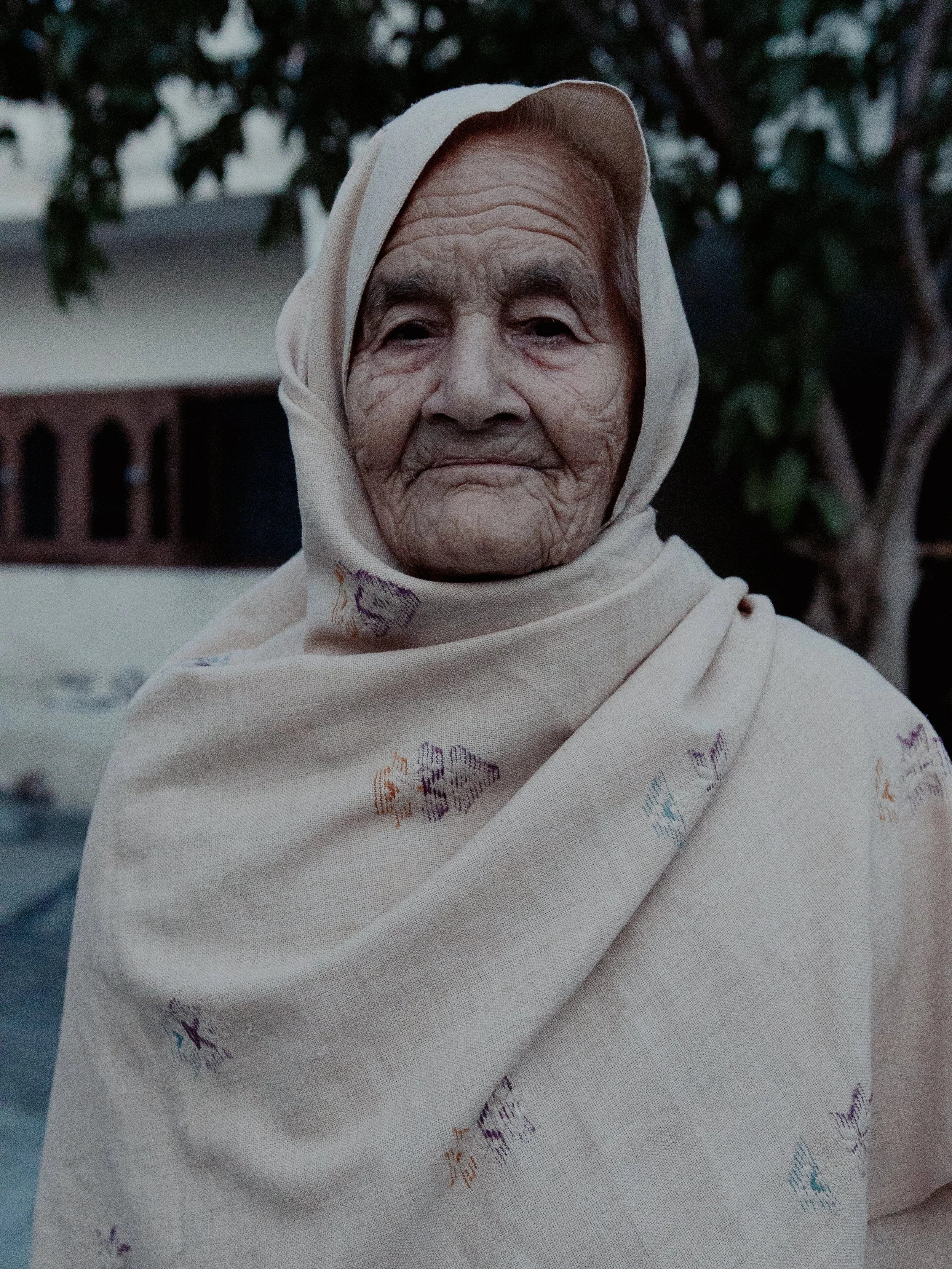 A close-up of an elderly woman with a gentle expression, wrapped in a cream-colored shawl with small, colorful embroidered patterns, standing outdoors with trees and a house in the background.