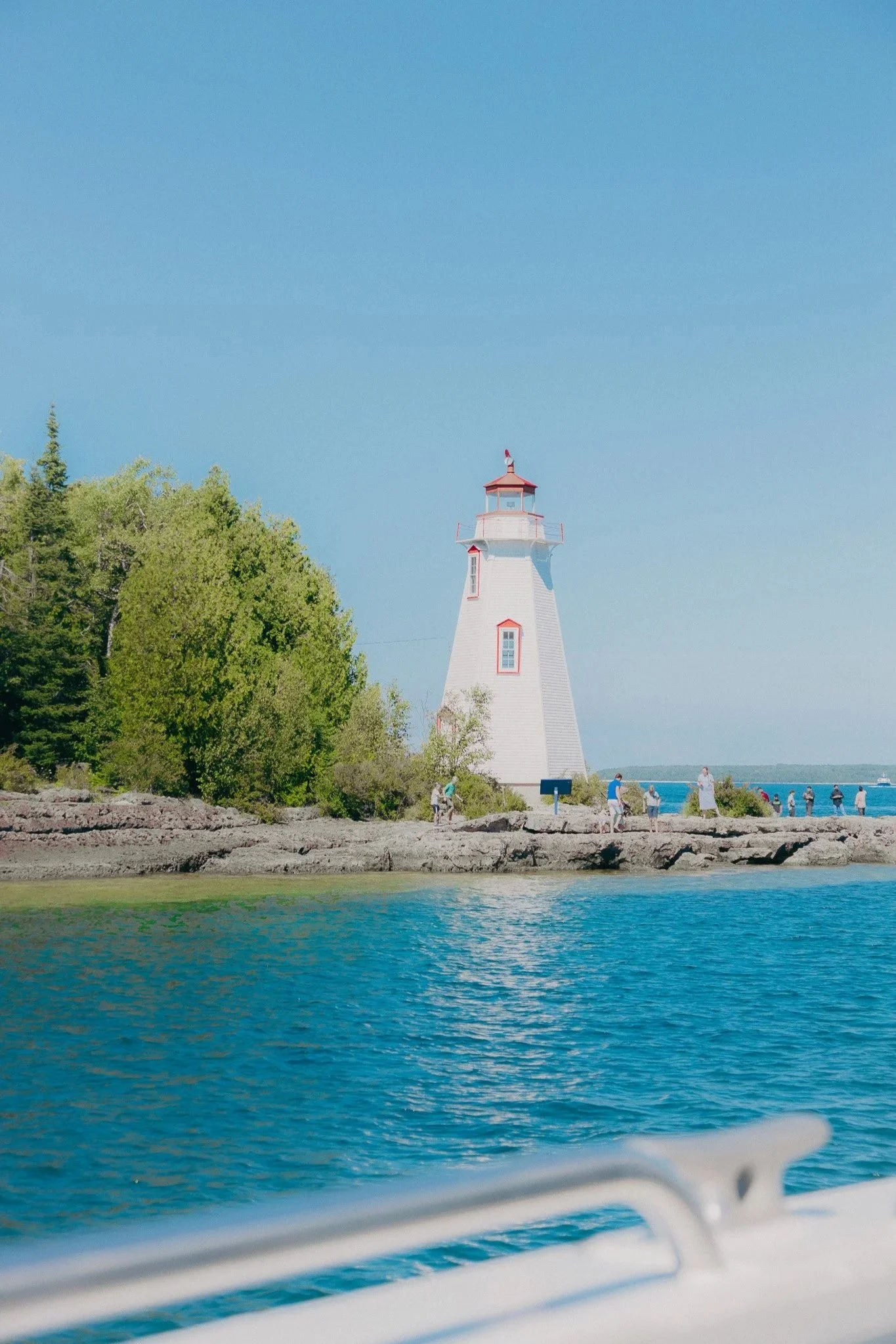 A white lighthouse with red trim on a rocky shoreline with trees, under a clear blue sky, near a body of water with a boat in the foreground.