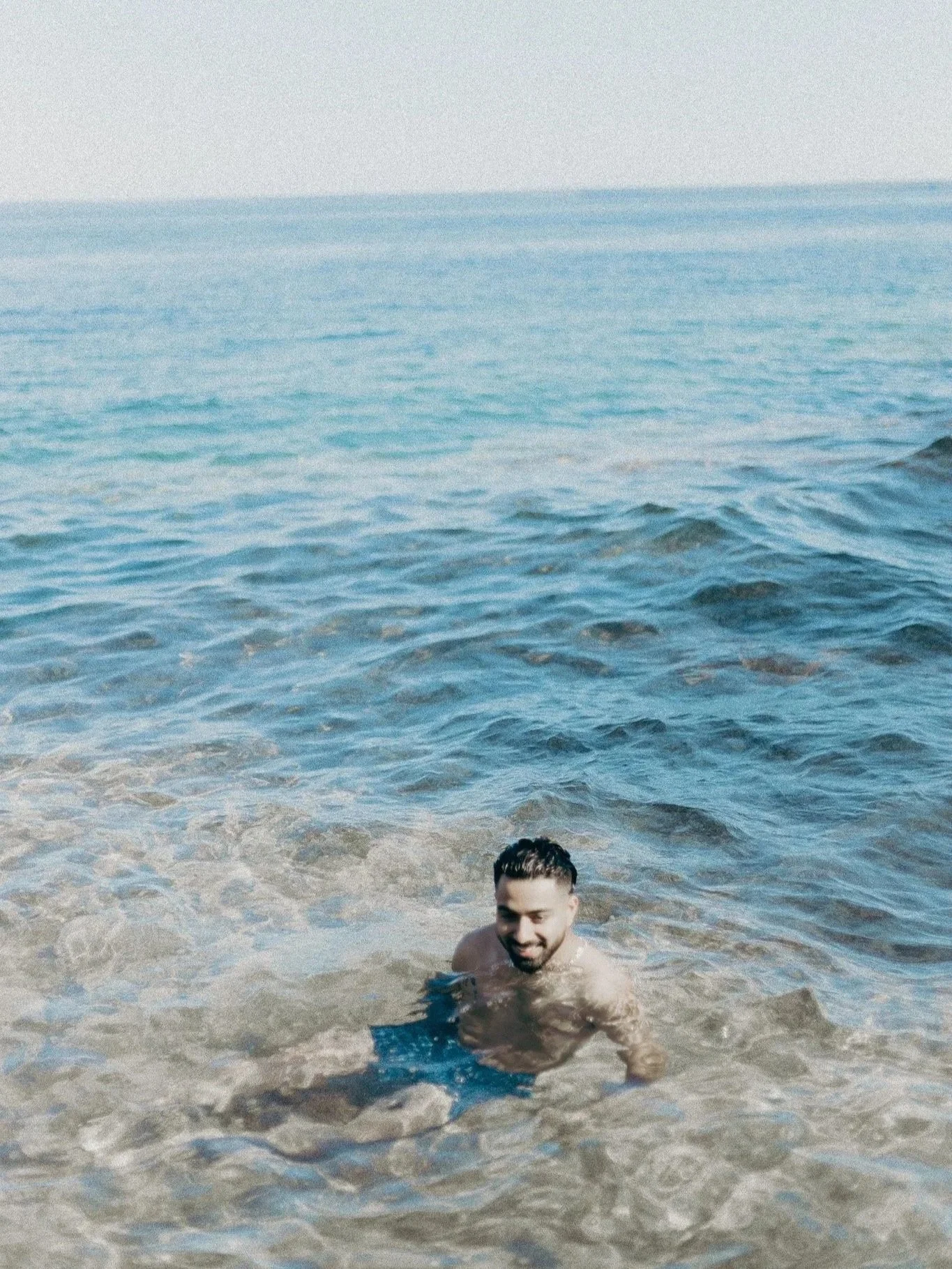 A smiling man with dark hair and a beard swimming in the ocean with clear blue water and gentle waves, under a mostly clear sky.
