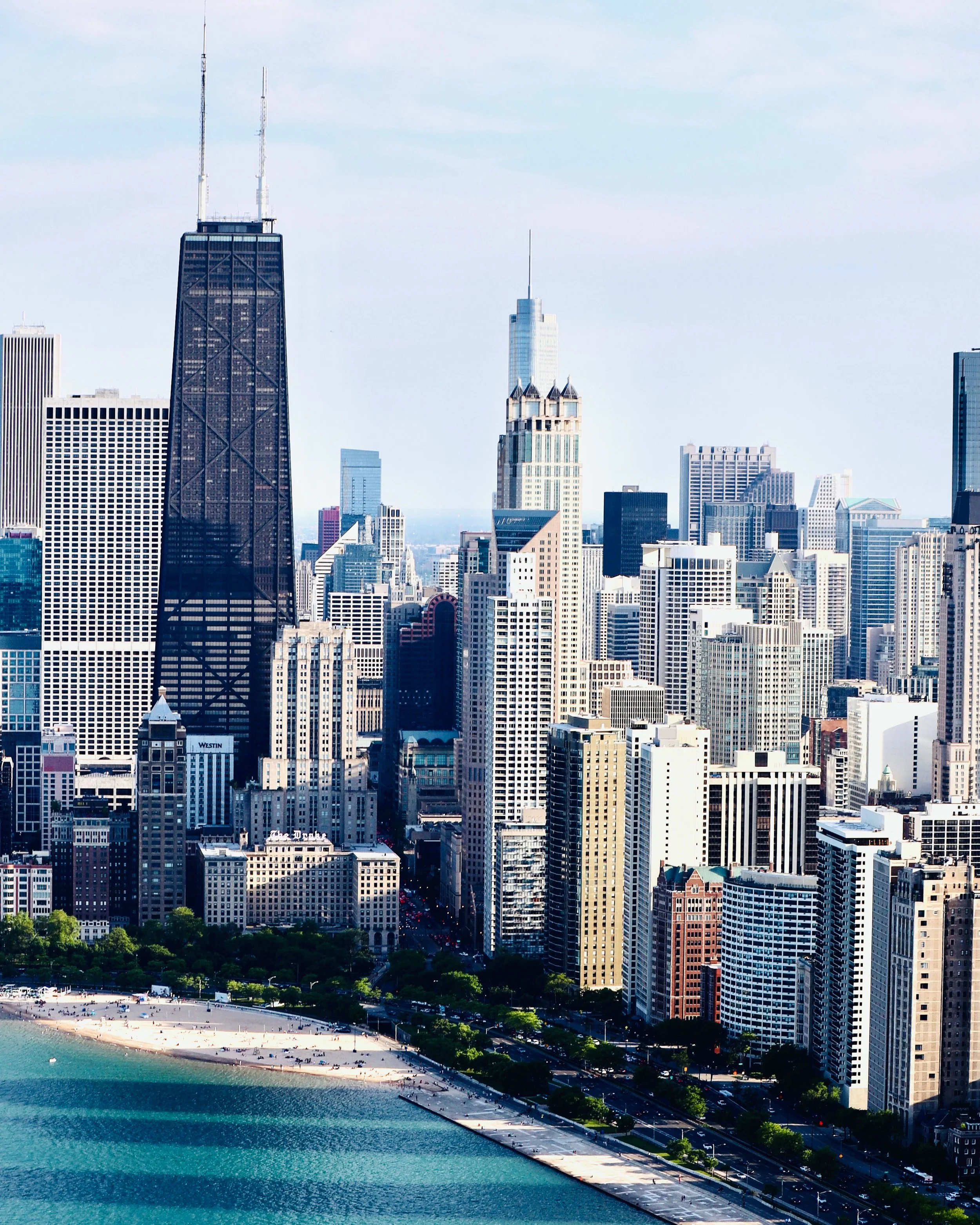 Skyline of Chicago with tall skyscrapers, Lake Michigan in the foreground, and a mostly clear sky.