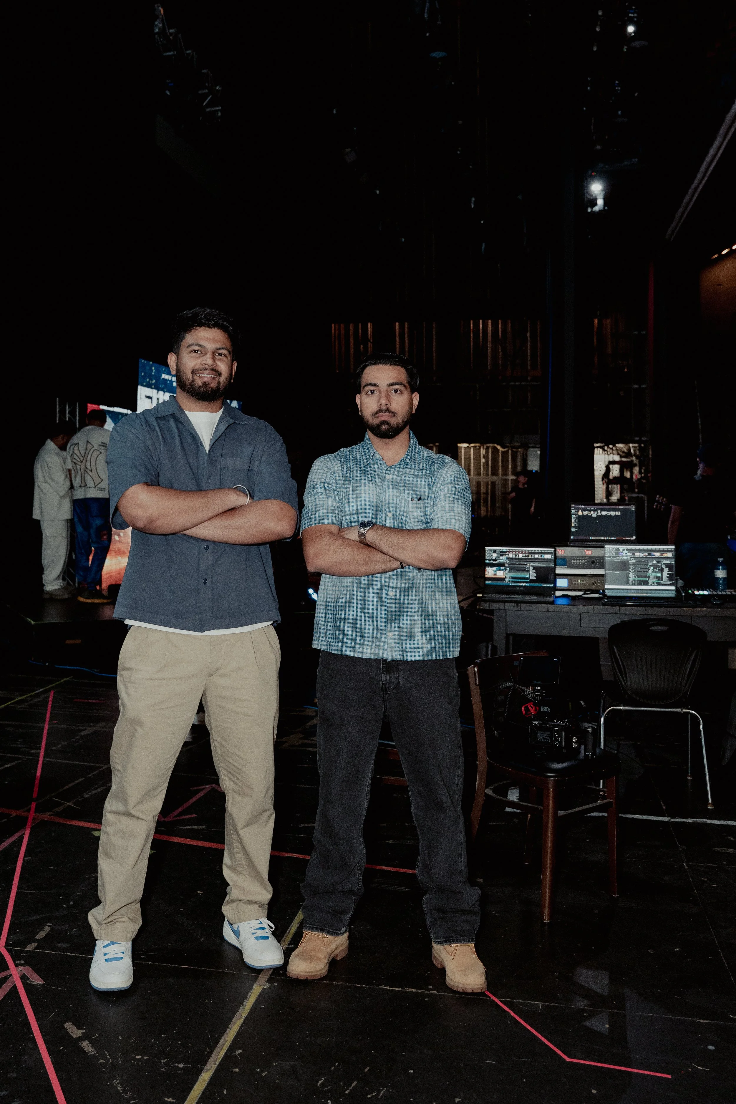 Two men standing with arms crossed in a dark indoor setting, with audio and lighting equipment visible behind them.