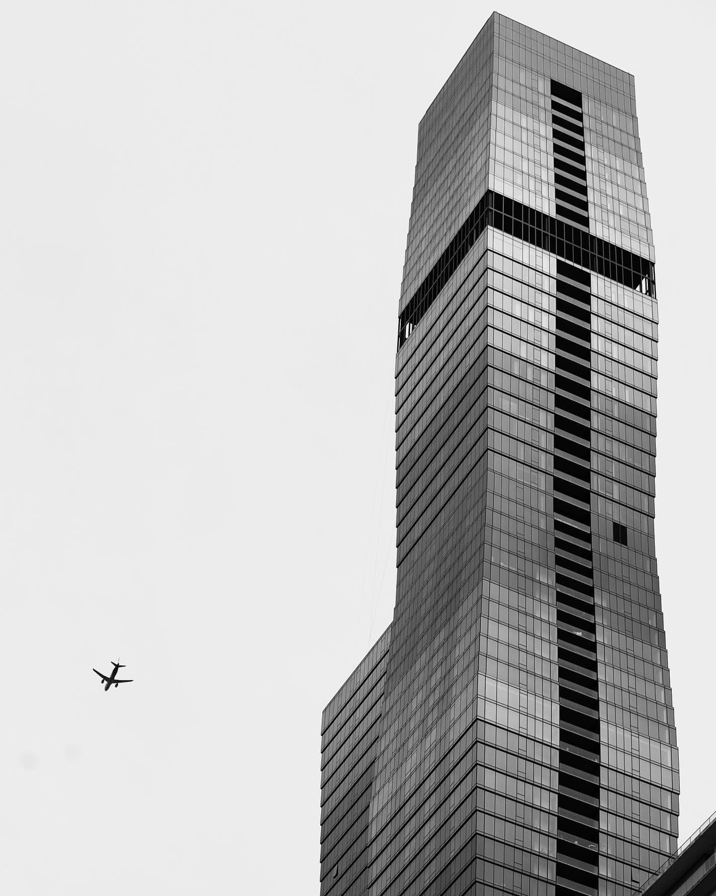 A tall modern glass skyscraper with geometric design and an airplane flying in the sky nearby.