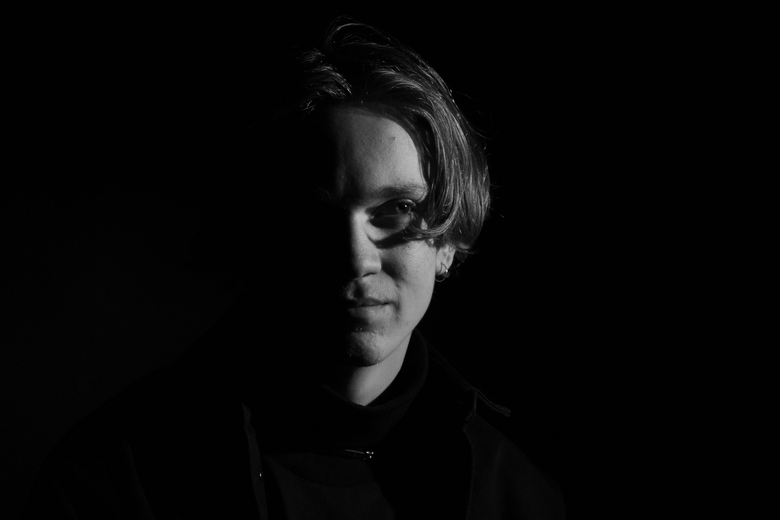 A black and white portrait of a young person with half of their face illuminated, showing their short, wavy hair and subtle facial features.