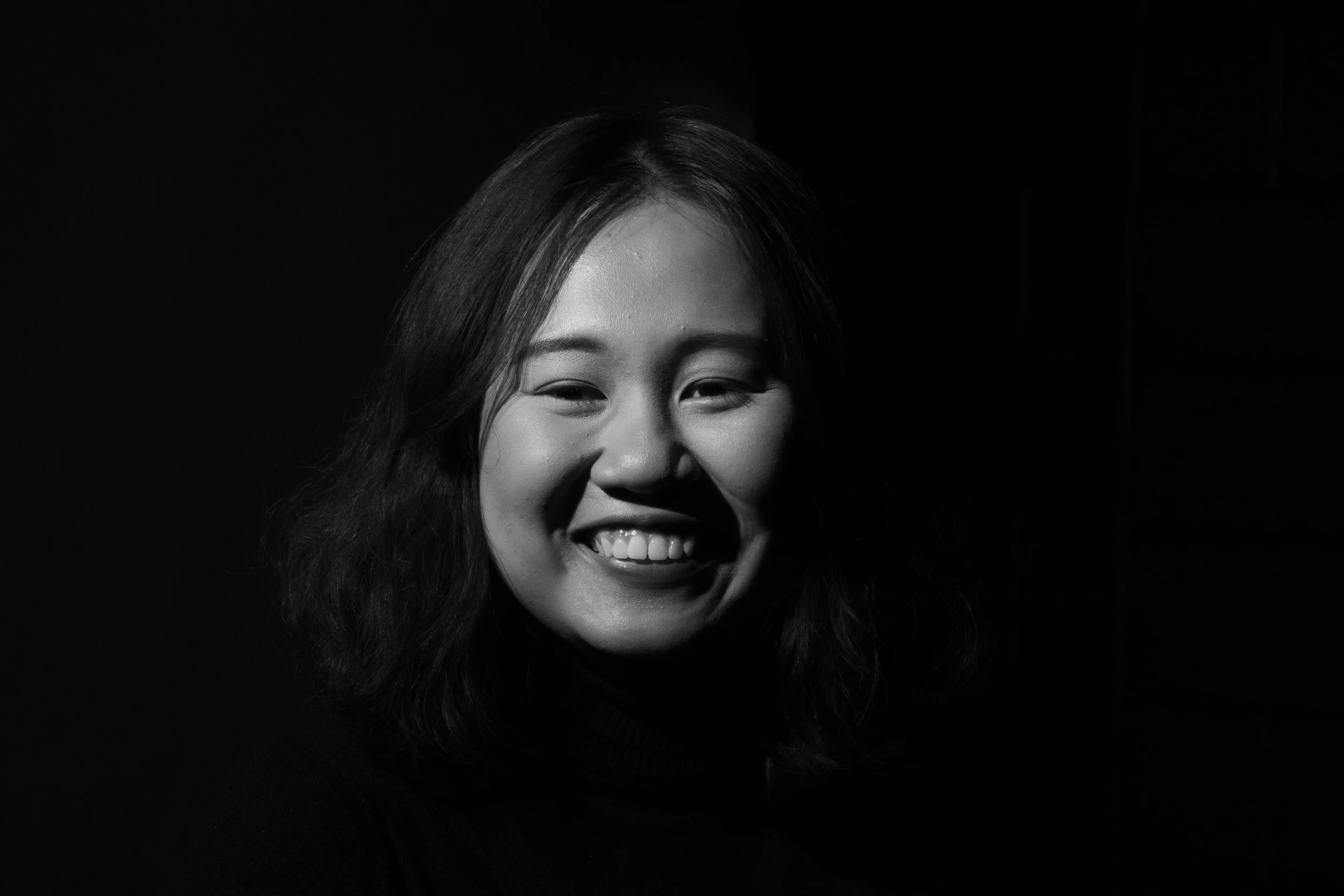 Black and white portrait of a smiling Asian woman with shoulder-length hair, wearing a dark turtleneck, against a dark background.