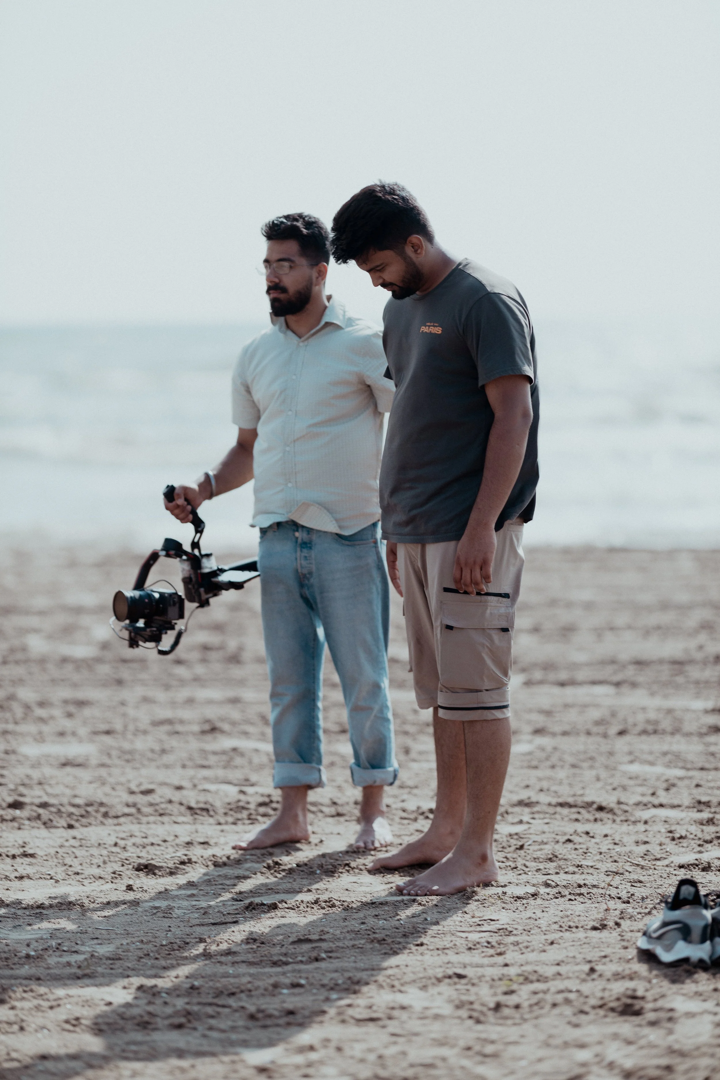 Two men standing barefoot on the sand at the beach, one holding a camera mounted on a stabilizer.