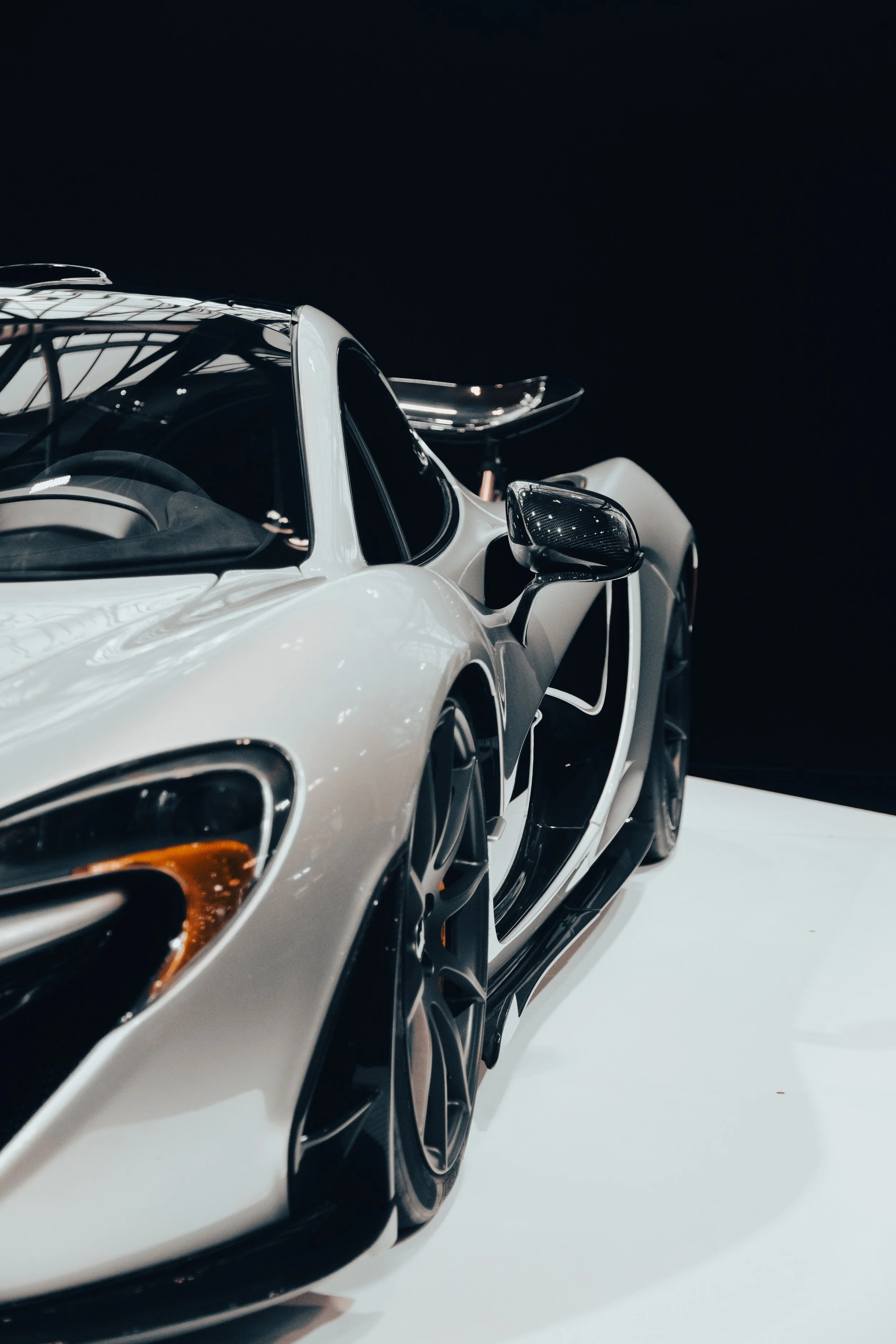 Close-up of a sleek, white sports car with black accents and a prominent rear wing, displayed on a white platform against a black background.