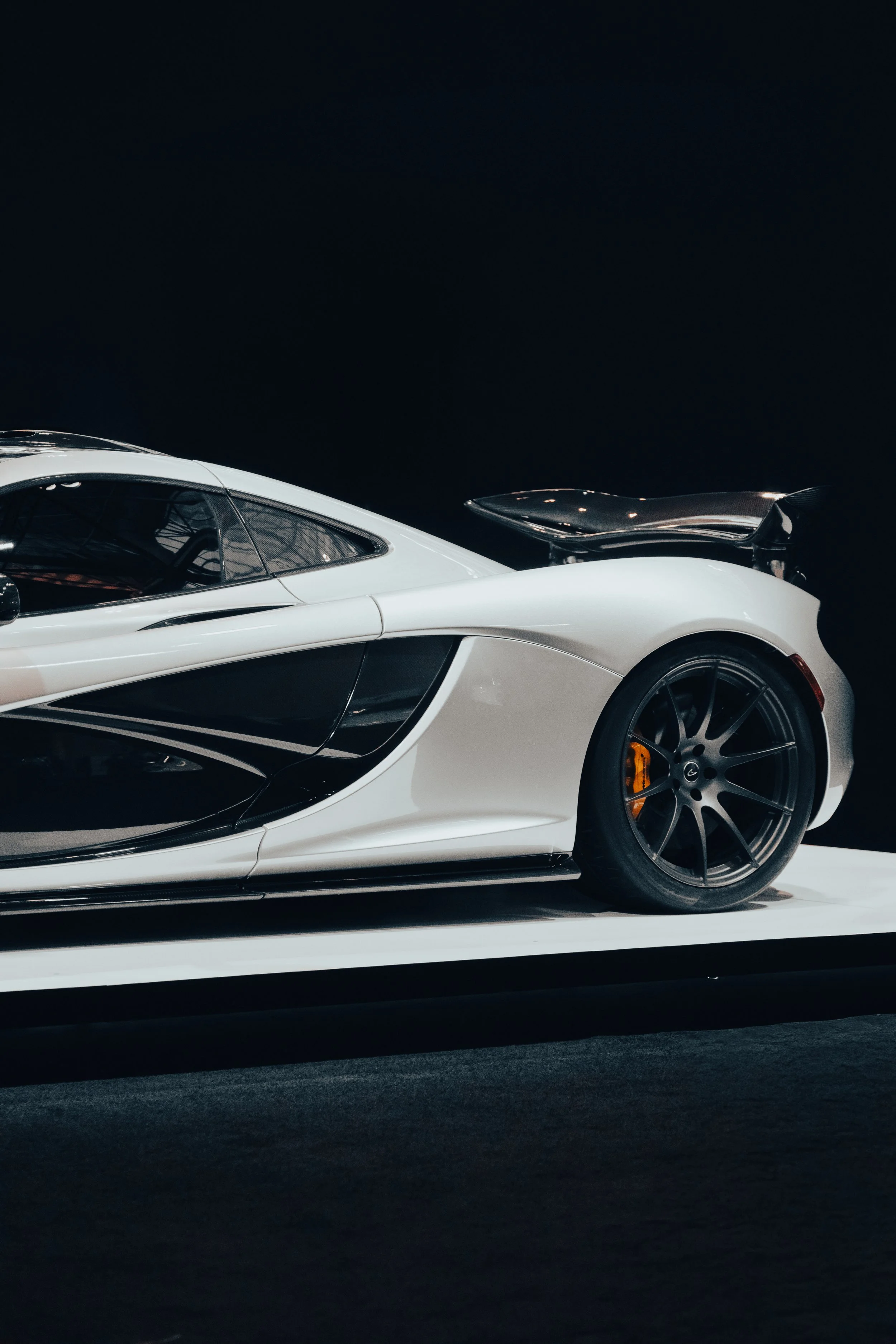 Side view of a sleek, white sports car with black wheels and a rear wing, displayed on a platform against a dark background.