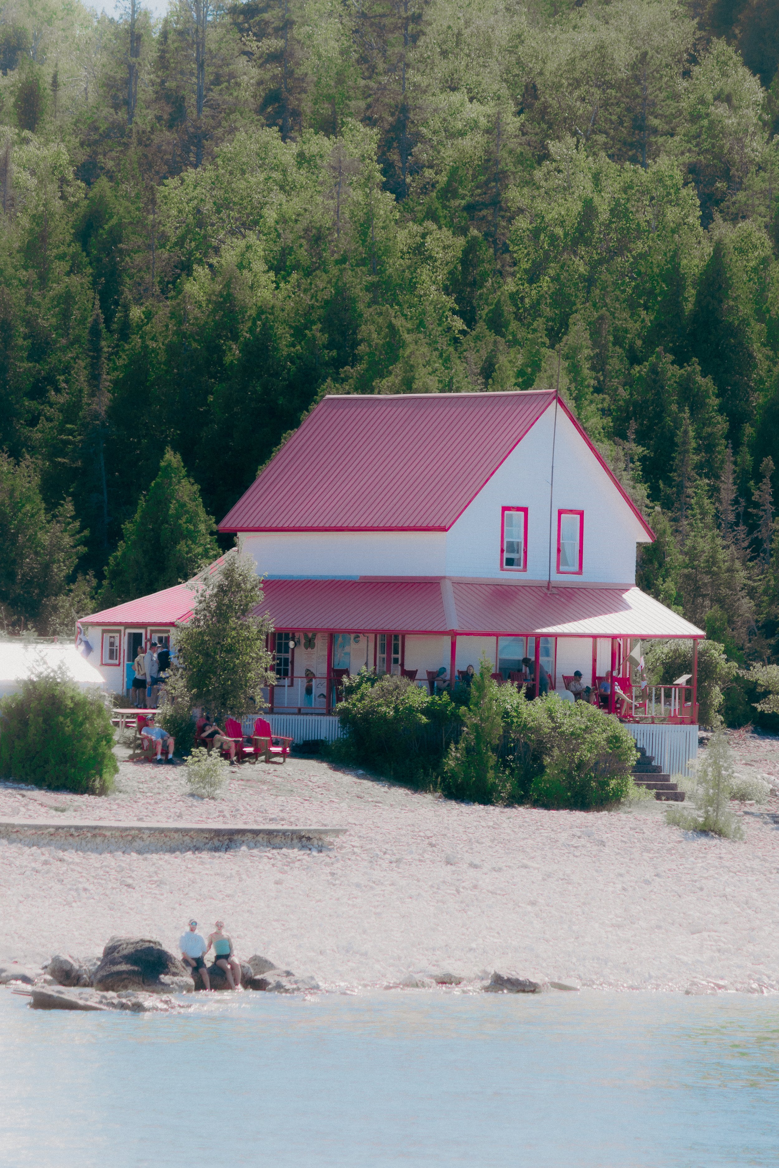 A two-story house with a pink roof and pink trim, situated on a sandy beach near a body of water, surrounded by trees. People are sitting outside on the porch and benches, with some by the water.