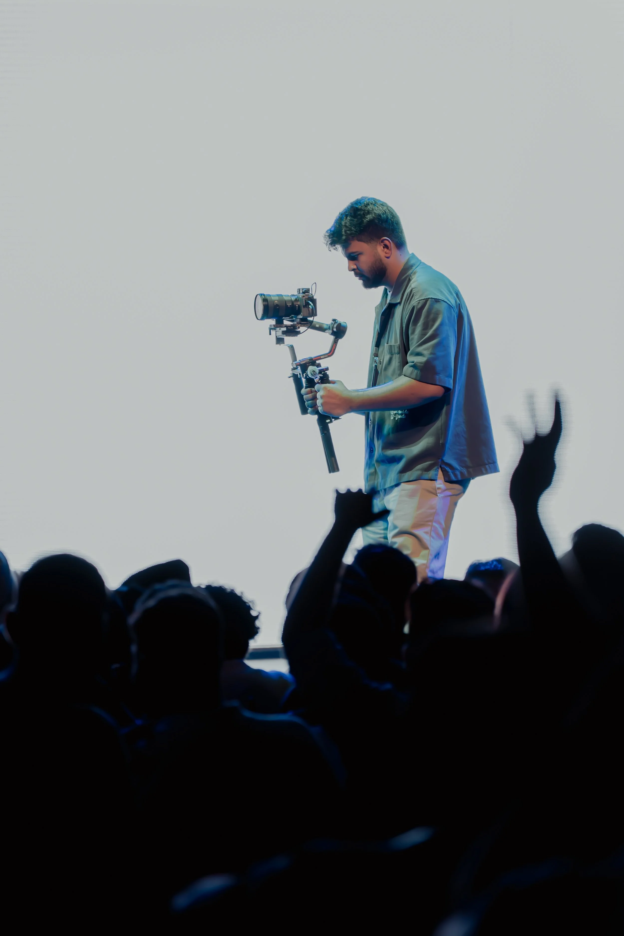 A young man operating a camera stabilizer and filming equipment during an event or performance, with an audience in the foreground.