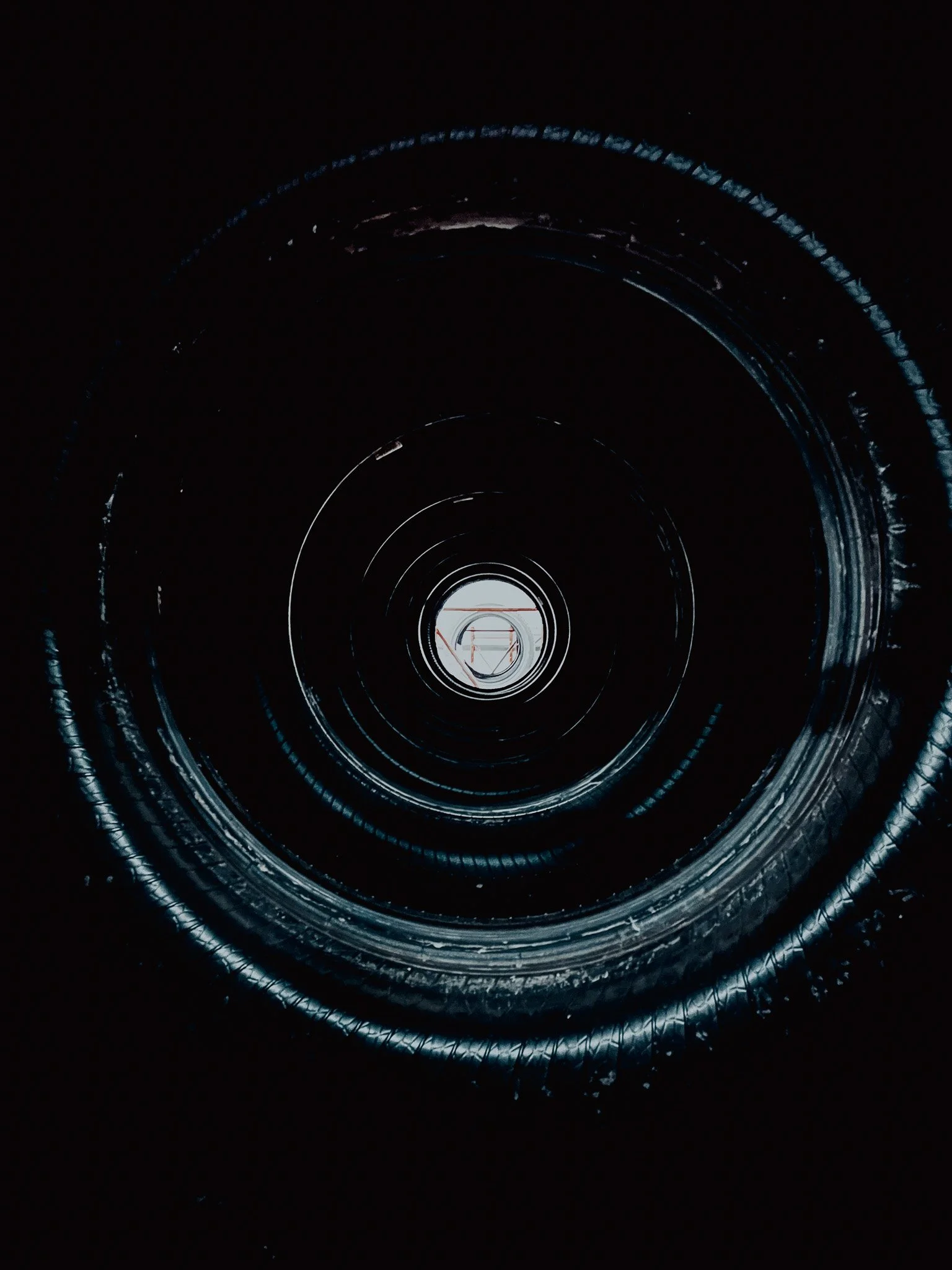 Looking inside a black cylindrical tunnel or pipe with a shiny interior, showing concentric rings and a small illuminated opening at the end.