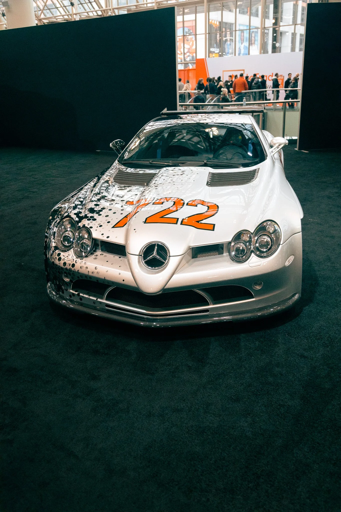 A silver Mercedes-Benz race car with the number 22 painted in orange on the hood, displayed on a dark green carpet at an indoor event with people in the background.
