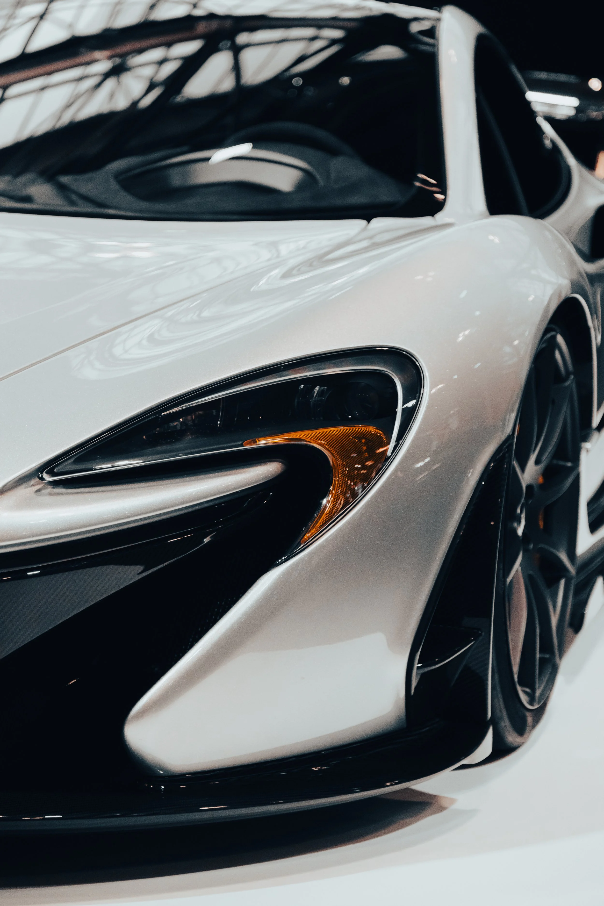 Close-up of a silver sports car's front left side, showing the headlight, hood, and part of the front wheel at an indoor car show.