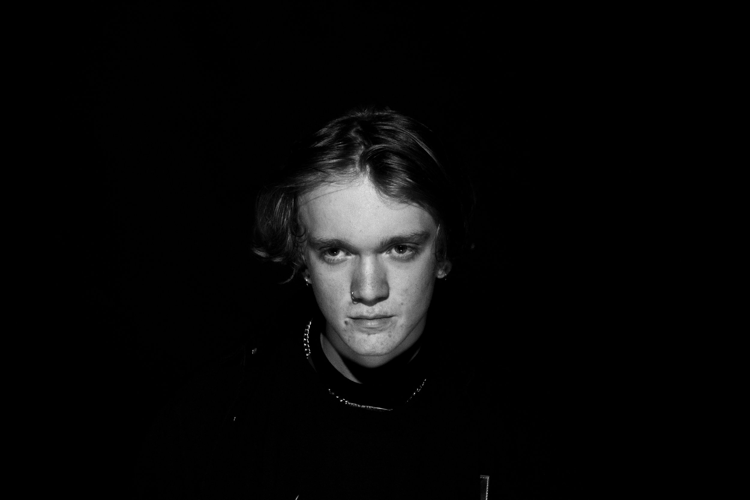 Black and white portrait of a young person with short hair, piercings, and a serious expression, centered against a dark background.