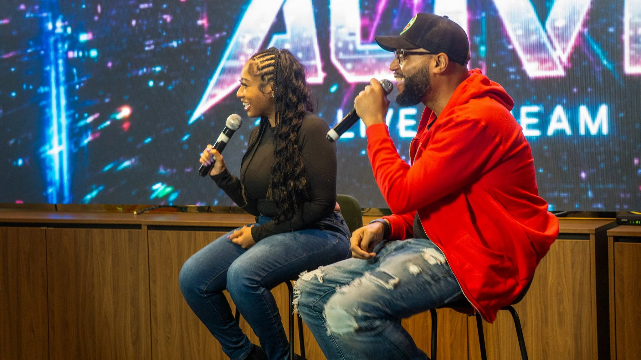 Two people are sitting on stools holding microphones on stage, smiling and speaking. Behind them, there is a large screen with neon-style graphics and the words 'LIVE STREAM' visible.