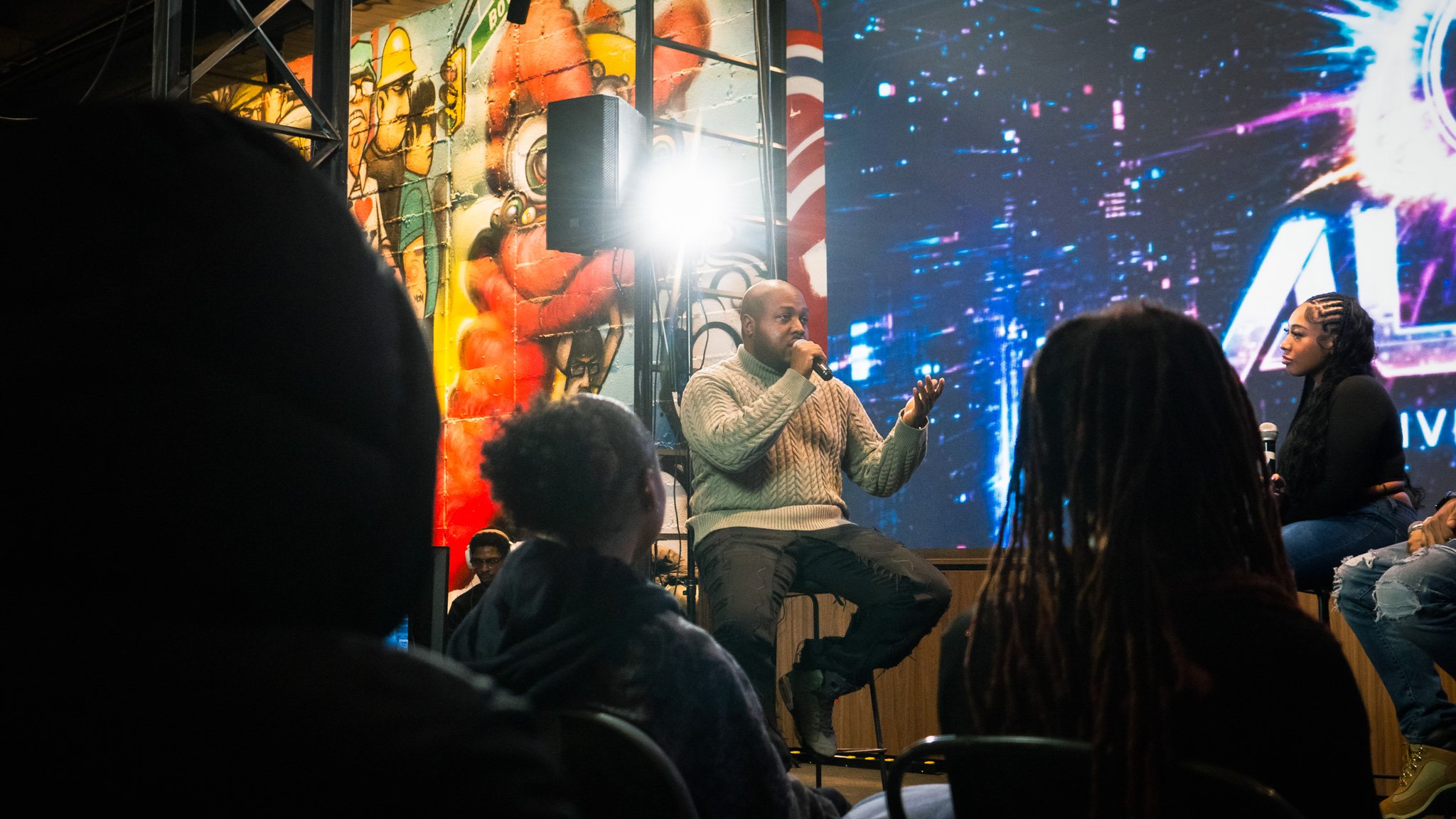 A man in a beige cable-knit sweater speaking into a microphone on a stage with a large screen and mural background, and a woman sitting beside him holding a microphone, with an audience watching.