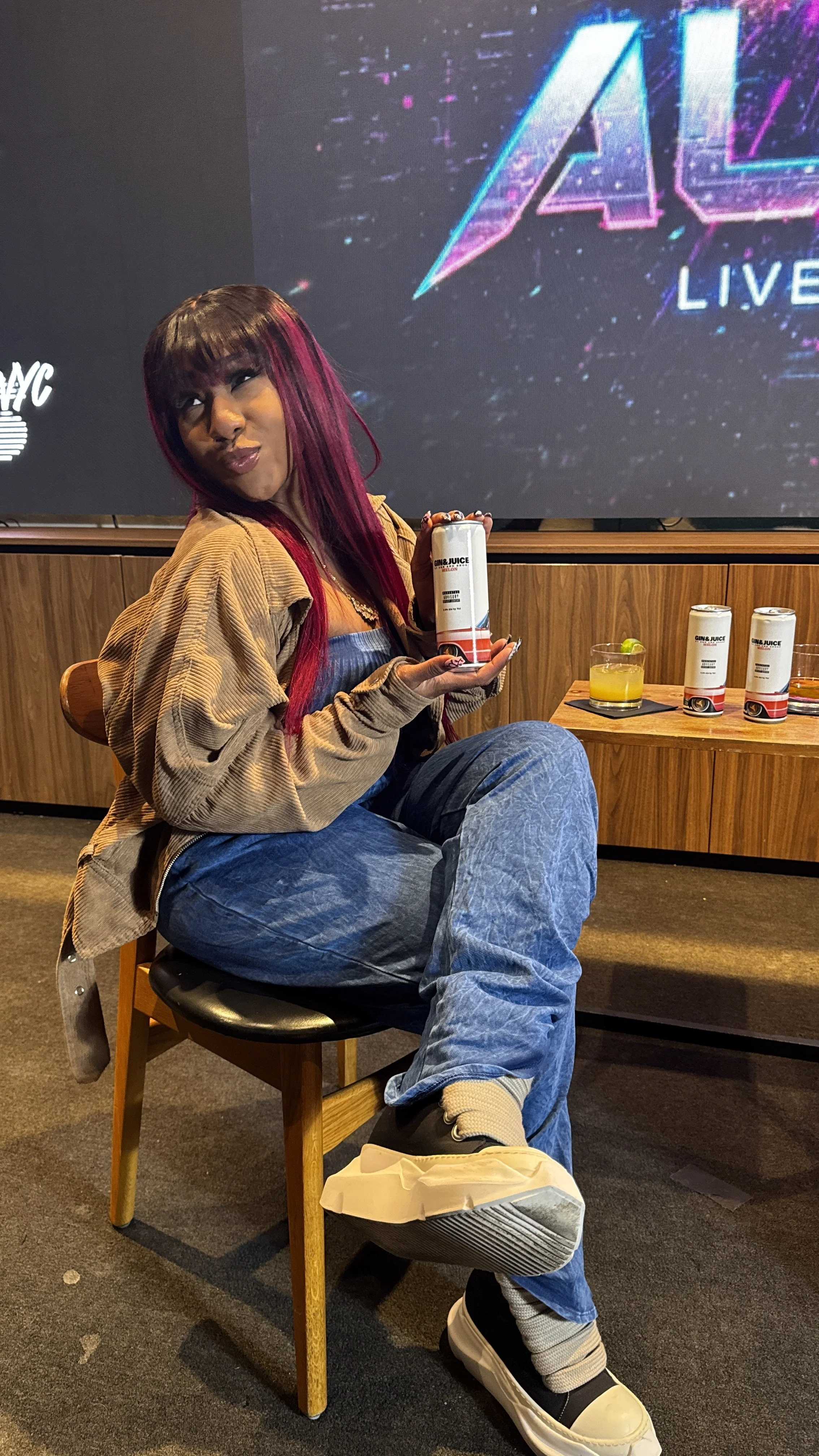 A woman with pink and black hair sitting on a wooden chair in front of a large screen, holding a can of energy drink. There are three more cans and two glasses with beverages on a wooden table beside her.