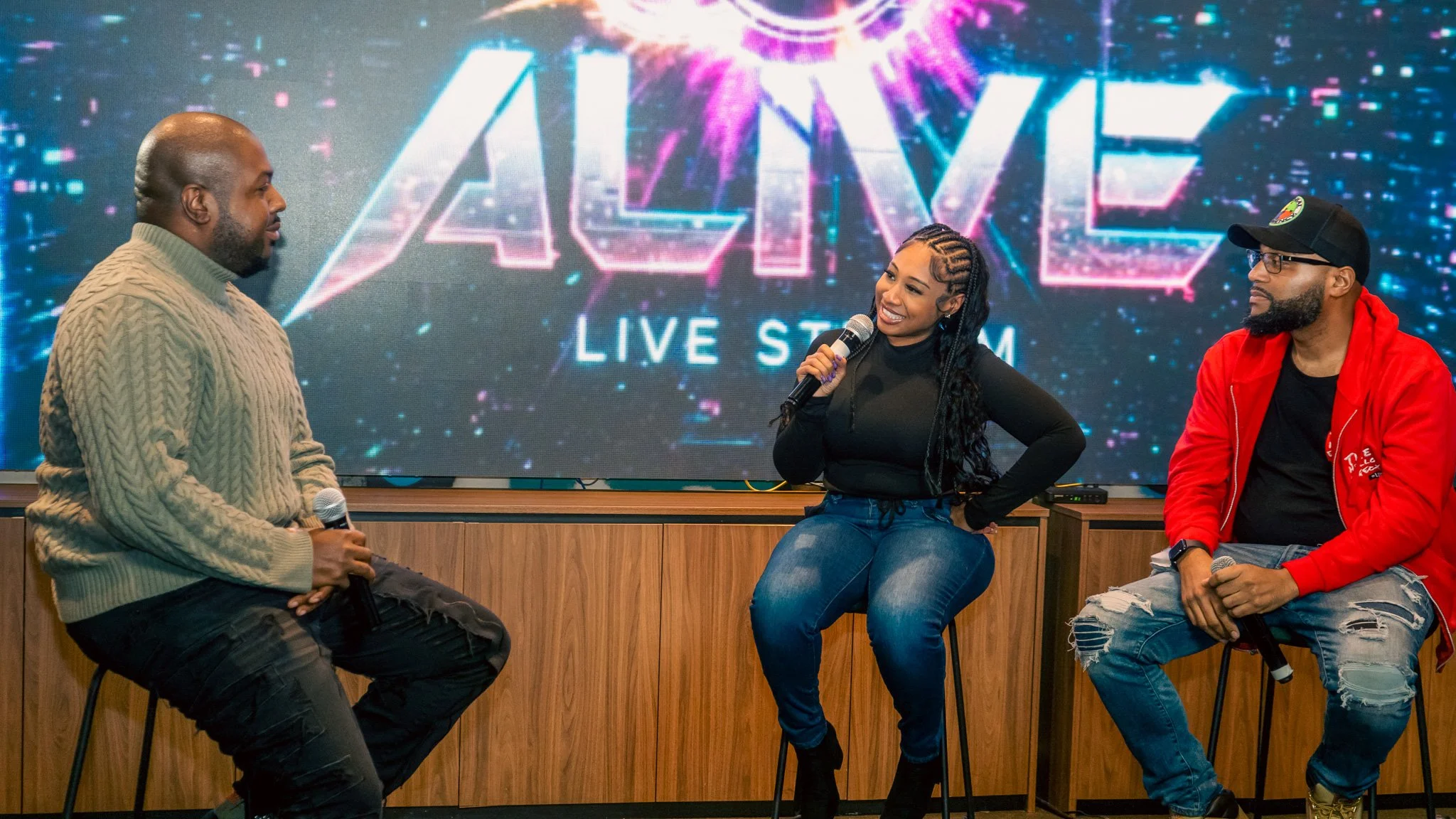 Three people sitting on stools in front of a large screen that says 'ALIVE LIVE STREAM'. They are participating in a panel or interview, with microphones in hand. The woman in the middle is smiling and speaking, wearing a black top and jeans, while t