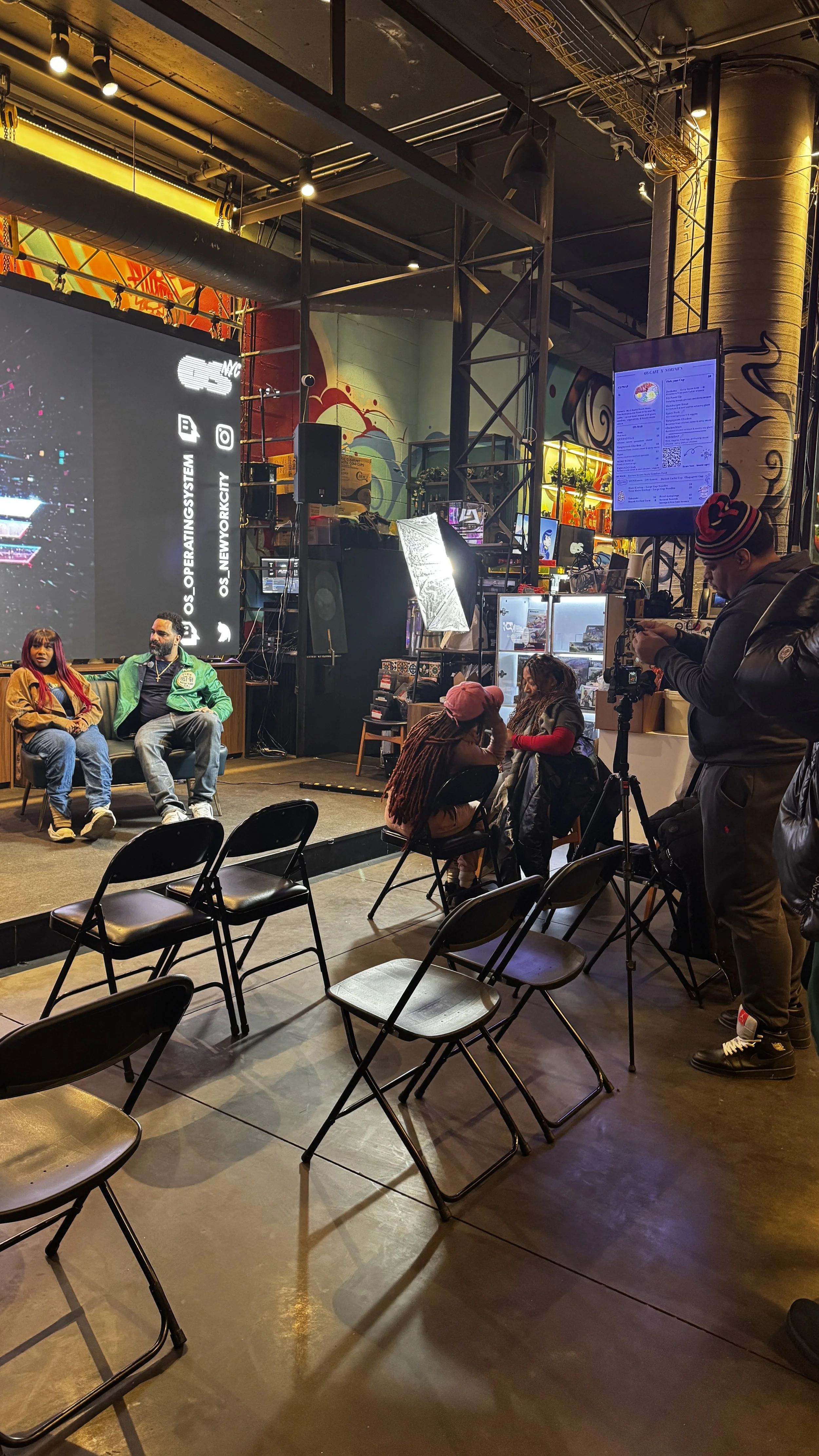 People sitting on a stage in front of a large screen, with a photographer and others capturing the event in a colorful, graffiti-decorated venue.
