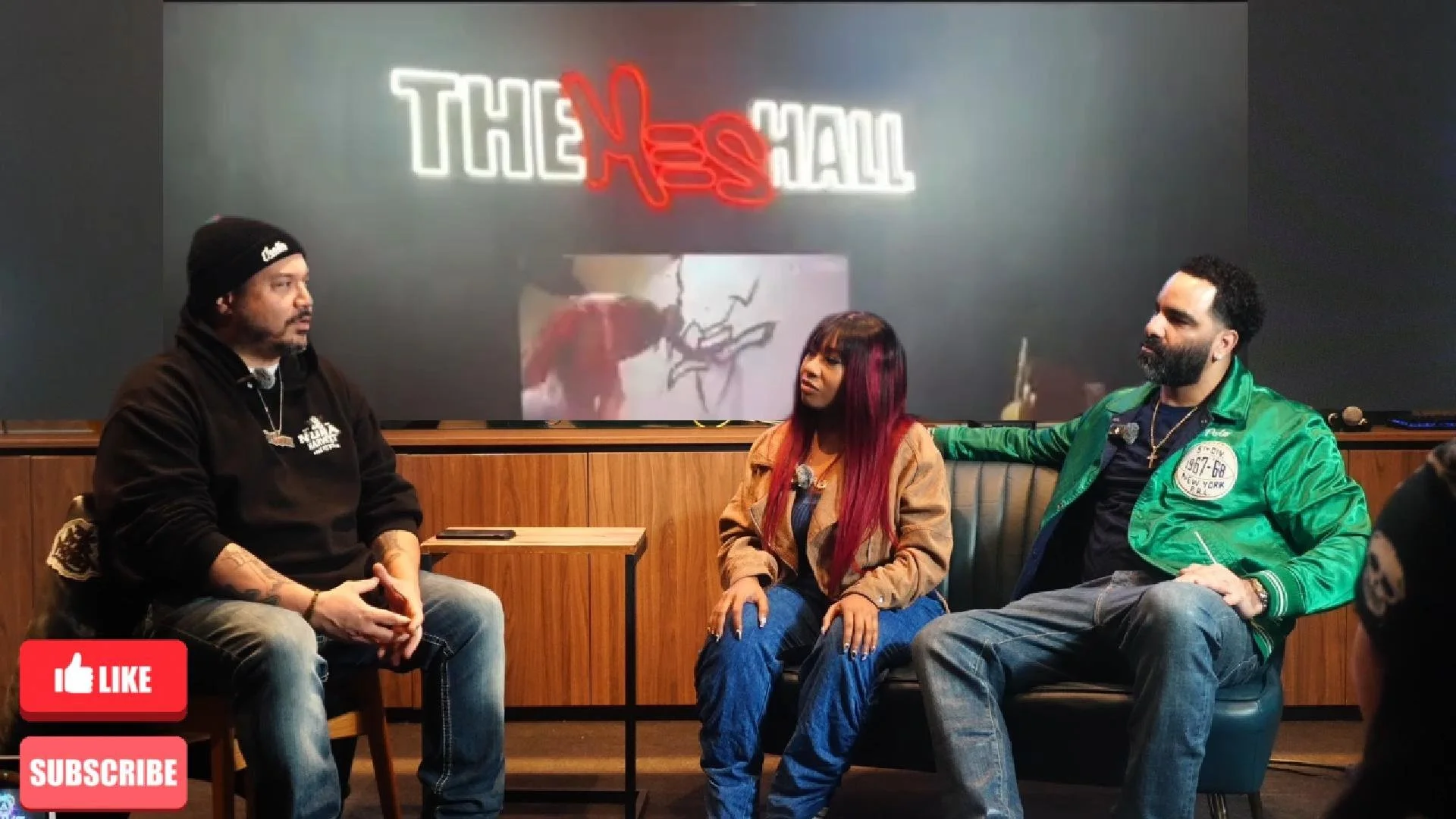 Three people sitting on a couch and a chair in a room, engaged in conversation. Behind them, a wall with a neon sign that reads 'THE HAL'. Red and white thumbs-up icons with the words 'LIKE' and 'SUBSCRIBE' are in the bottom left corner.