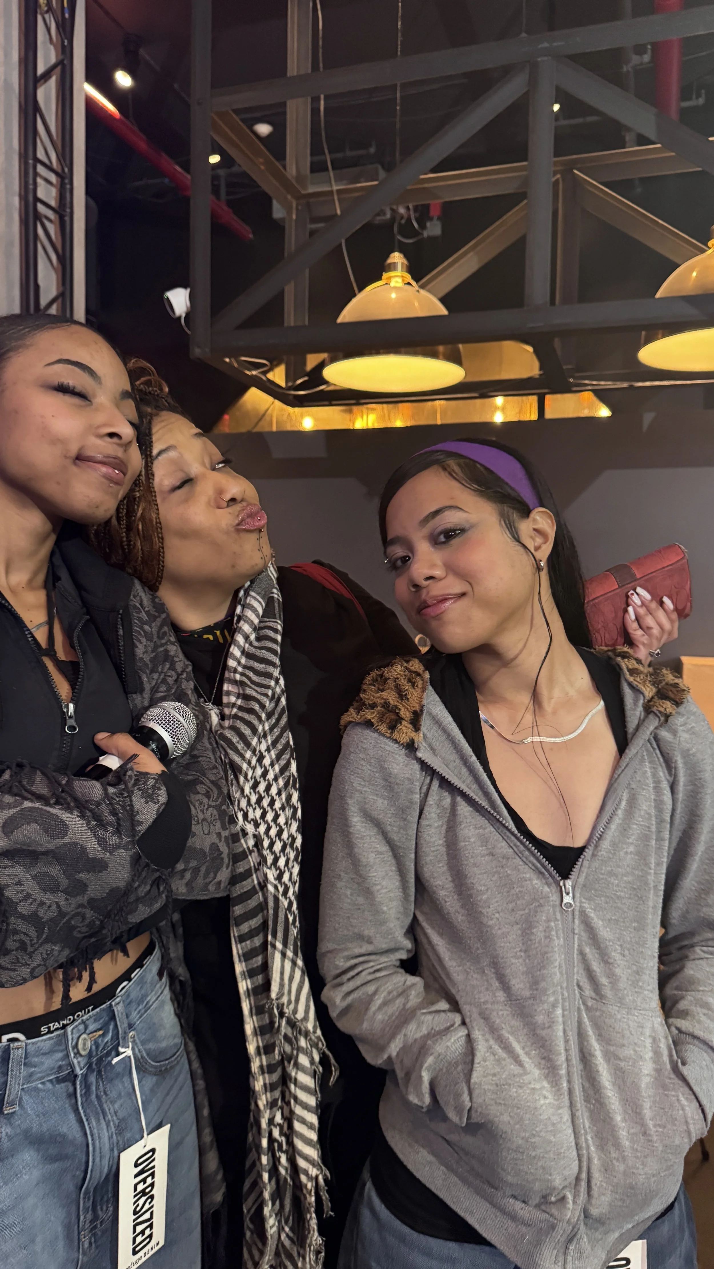 Three young women posing together indoors. One woman on the left is smiling with eyes closed, the middle woman is puckering lips, and the woman on the right is smiling slightly, wearing a hoodie and headphones. There are hanging lights and ceiling st