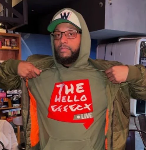 A man wearing glasses and a cap with a 'W' logo, pulling open his hoodie to reveal a shirt with the text 'THE HELLO EFFECT LIVE' in red and white.
