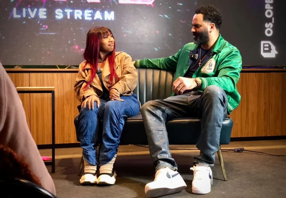 A woman and a man sitting on a black bench on a stage, engaging in a conversation. The woman has long red hair, a tan jacket, and jeans. The man has a beard, a green jacket, and jeans. There is a large screen behind them that says 'LIVE STREAM' and a