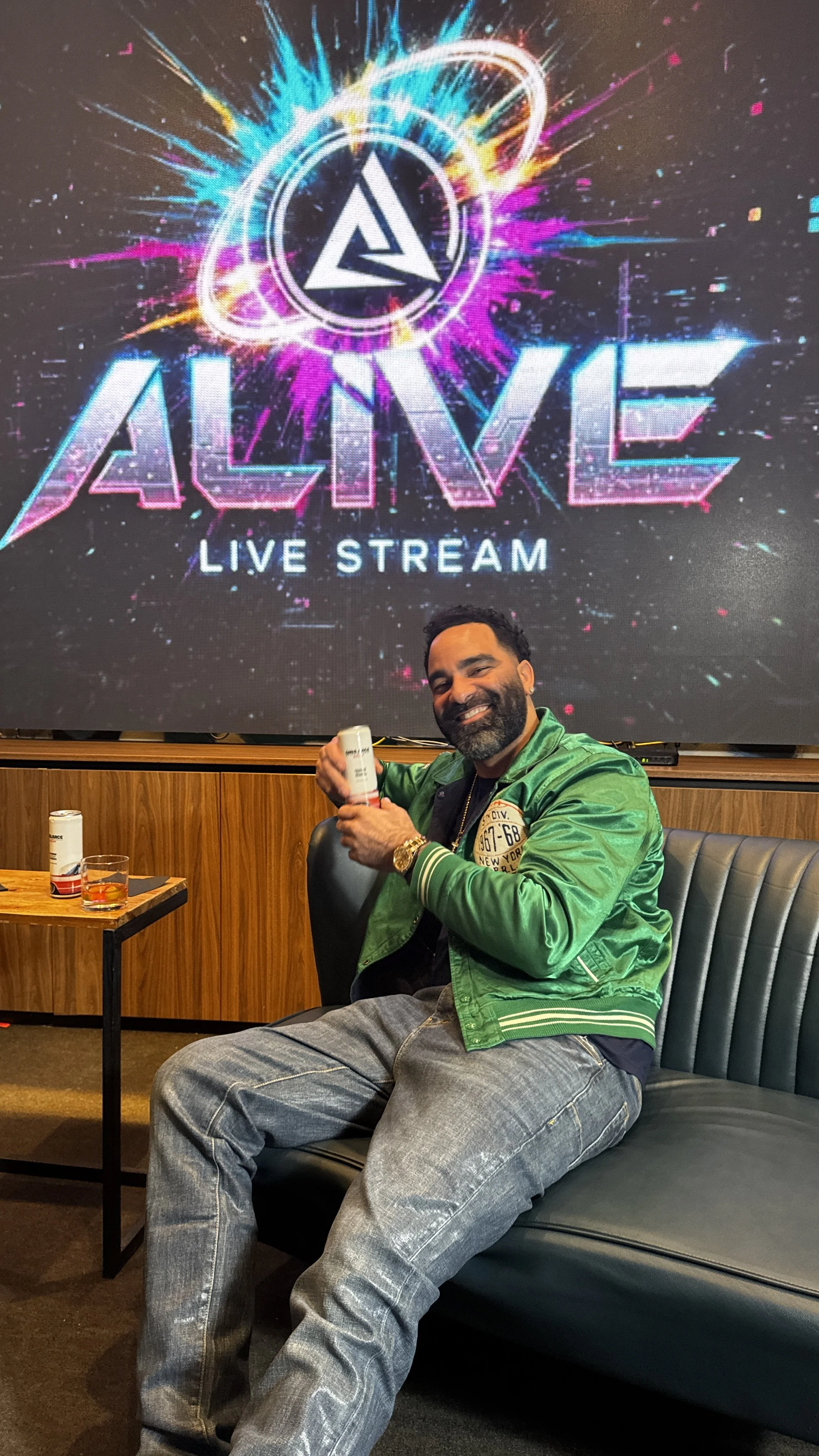 A man with a beard wearing a green jacket, seated on a black leather couch, smiling and holding a white bottle in his hand. Behind him is a large digital screen with a colorful logo and the words 'ALIVE LIVE STREAM.' There is a small table with a gla