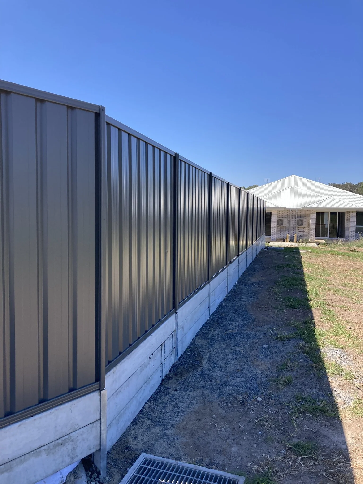 Side view of a dark gray metal fence with vertical panels, installed on a concrete base, beside a grassy area and a house with a white roof.