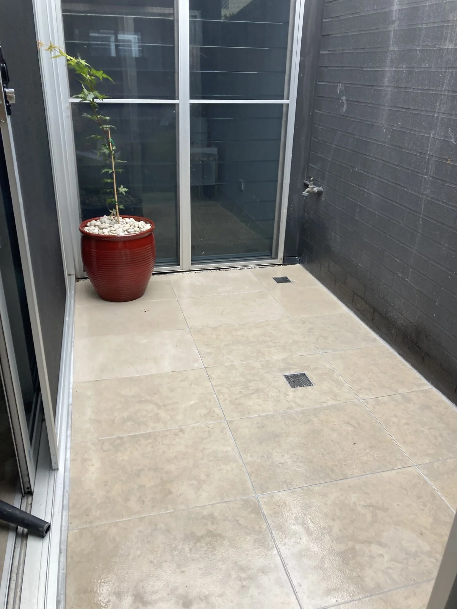 Small patio with beige tiles, a black brick wall, a glass sliding door, a water tap on the wall, and a red flower pot with a tall plant in it.