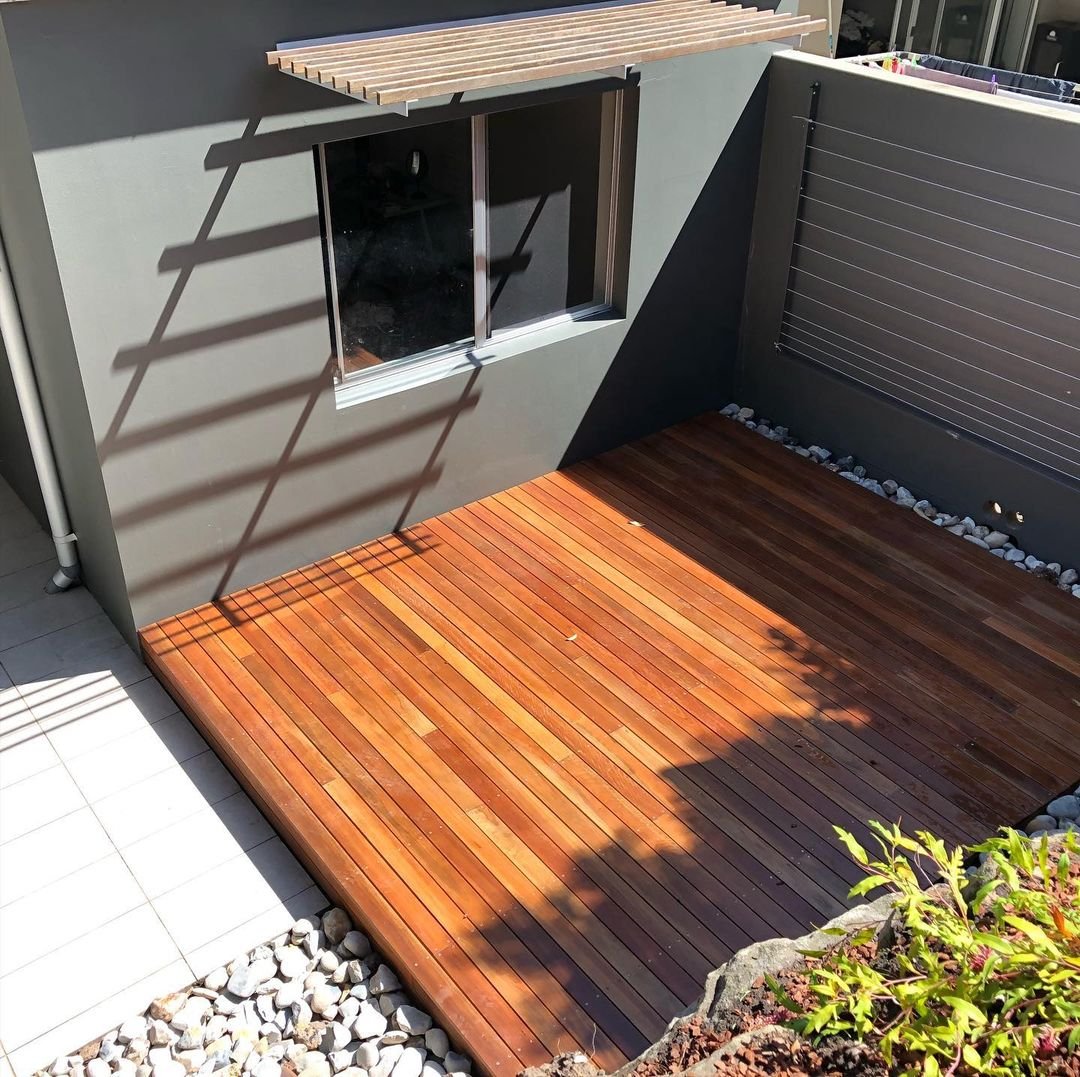 Small outdoor patio with wooden decking, a window with a wooden slatted sunshade, a privacy fence, and decorative rocks along the edges.