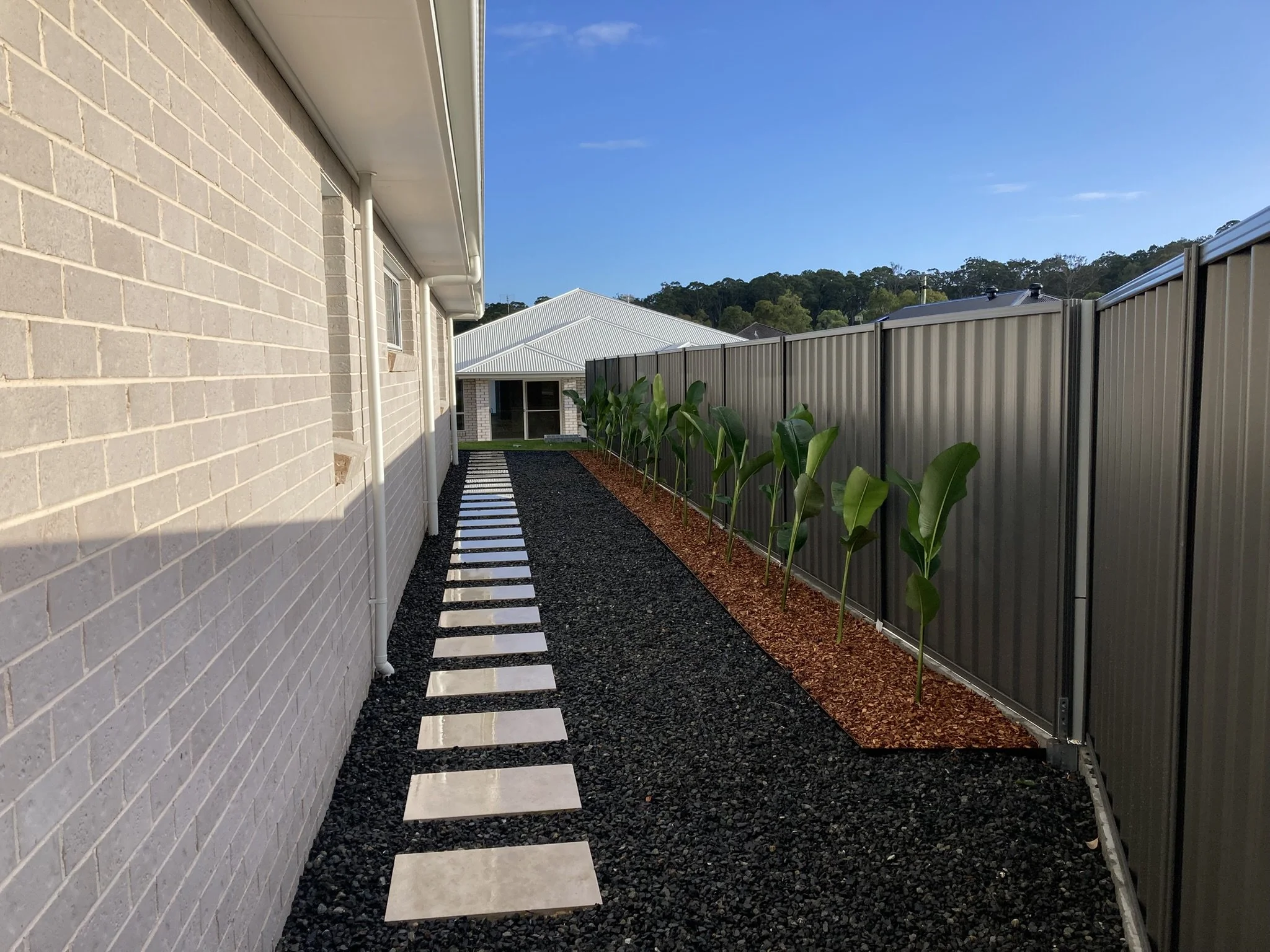 A narrow pathway with white stepping stones along a brick house wall on the left, and a row of young green plants on the right, separated by mulch. A grey metal fence runs parallel on the right, and a house with a metal roof is in the background under a blue sky.