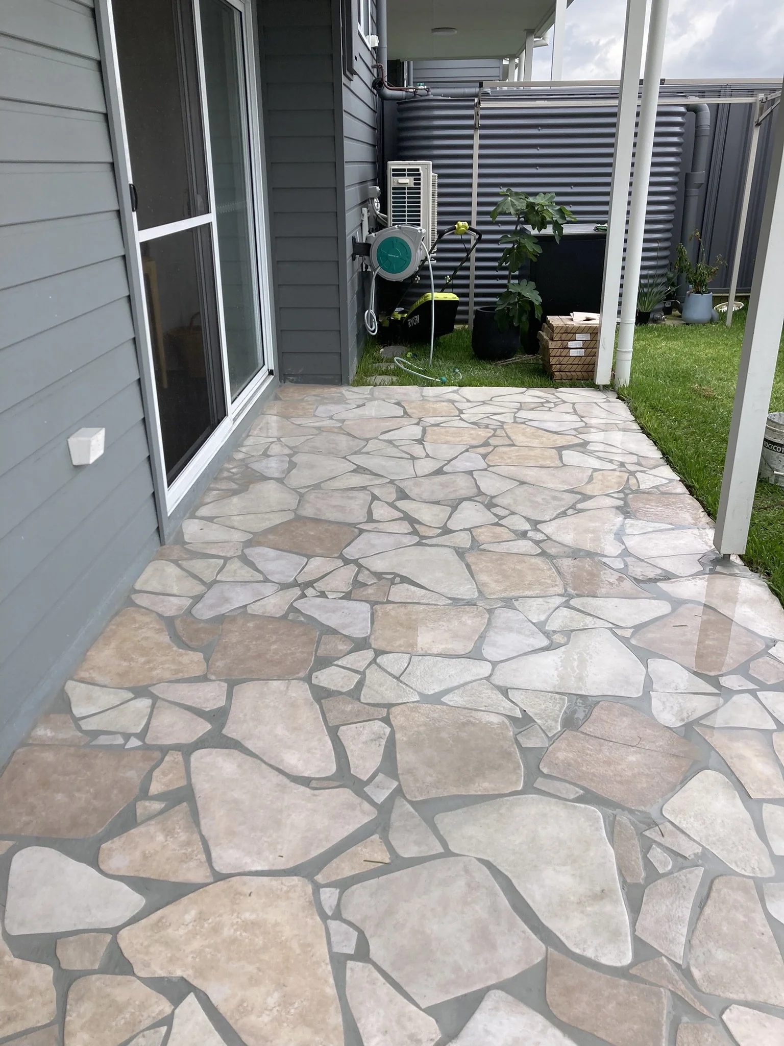 A backyard patio with a stone tile floor, grey siding house wall with a sliding glass door, and a fenced grassy yard area with potted plants and gardening tools.
