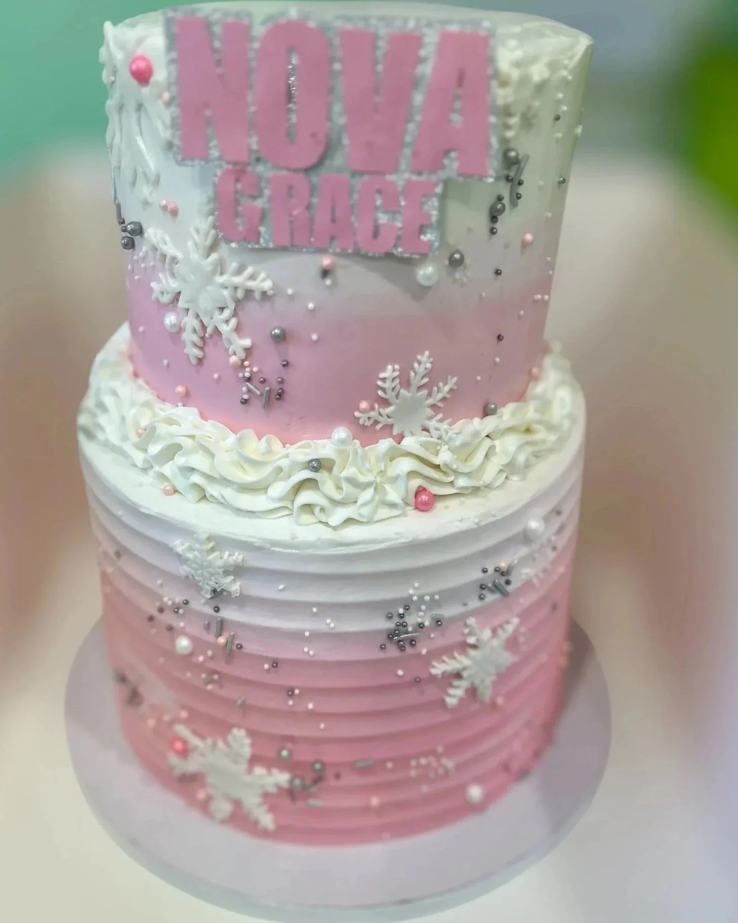 Two-tiered pink and white cake decorated with snowflakes, pearls, and silver beads, with a sign on the top reading "NOVA GRACE" in pink letters.